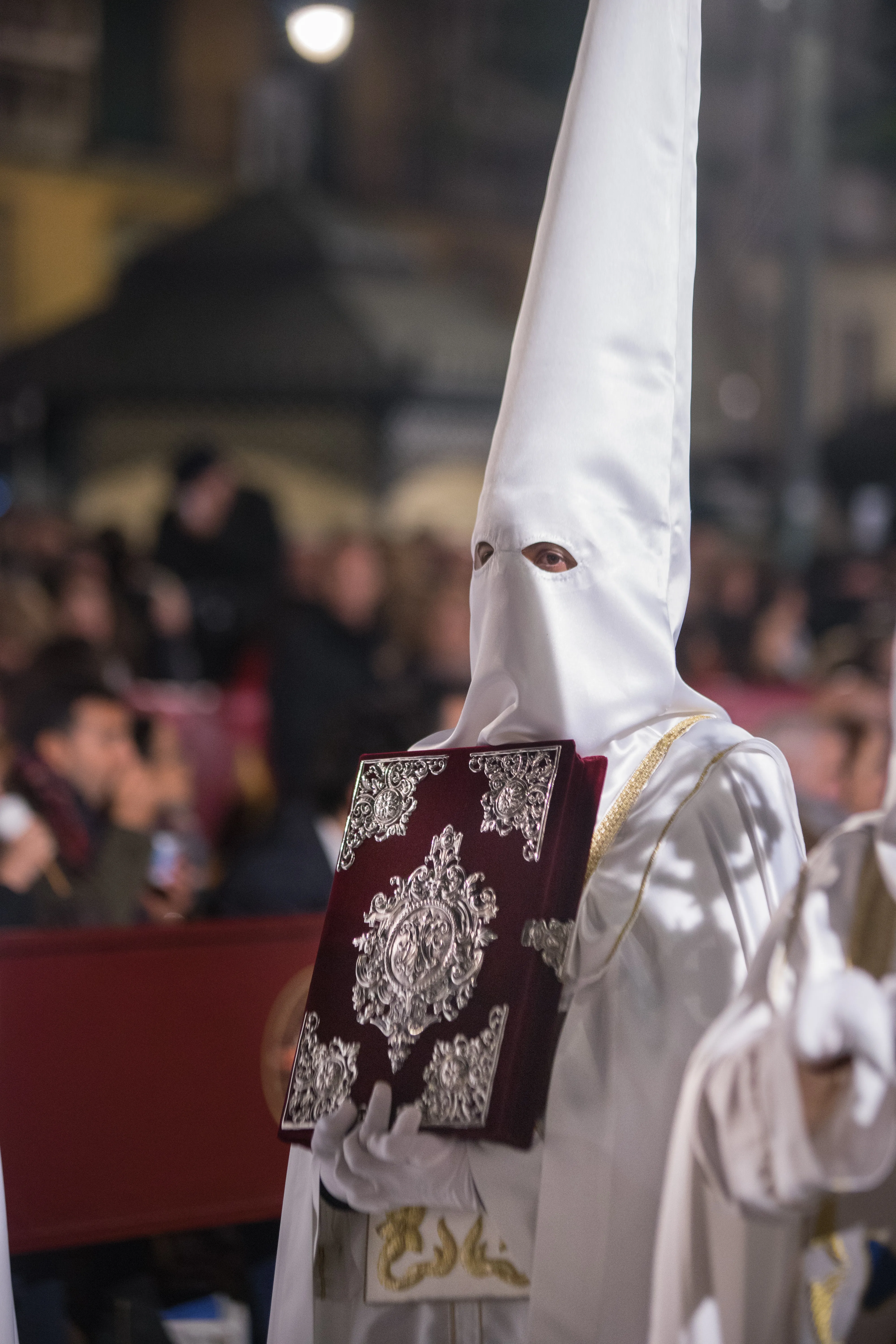 Un penitente con un alto capirote blanco sostiene un ornamentado libro ceremonial color granate con decoraciones de filigrana de plata durante una procesión nocturna de Semana Santa, con espectadores desenfocados al fondo.