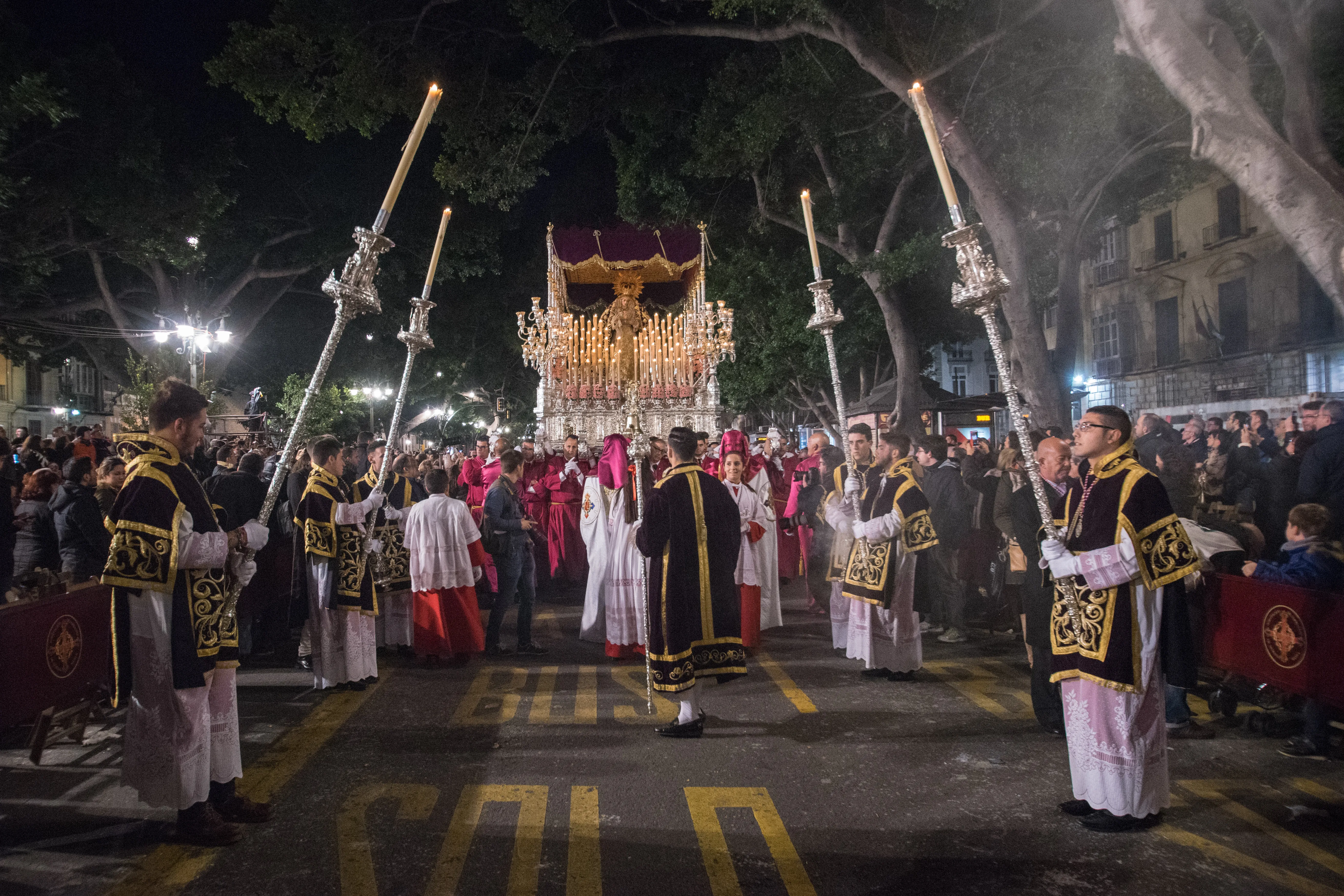 Procesión religiosa nocturna con miembros de la cofradía vestidos con túnicas y portando varas de plata, con un paso procesional iluminado al fondo en una calle de la ciudad.