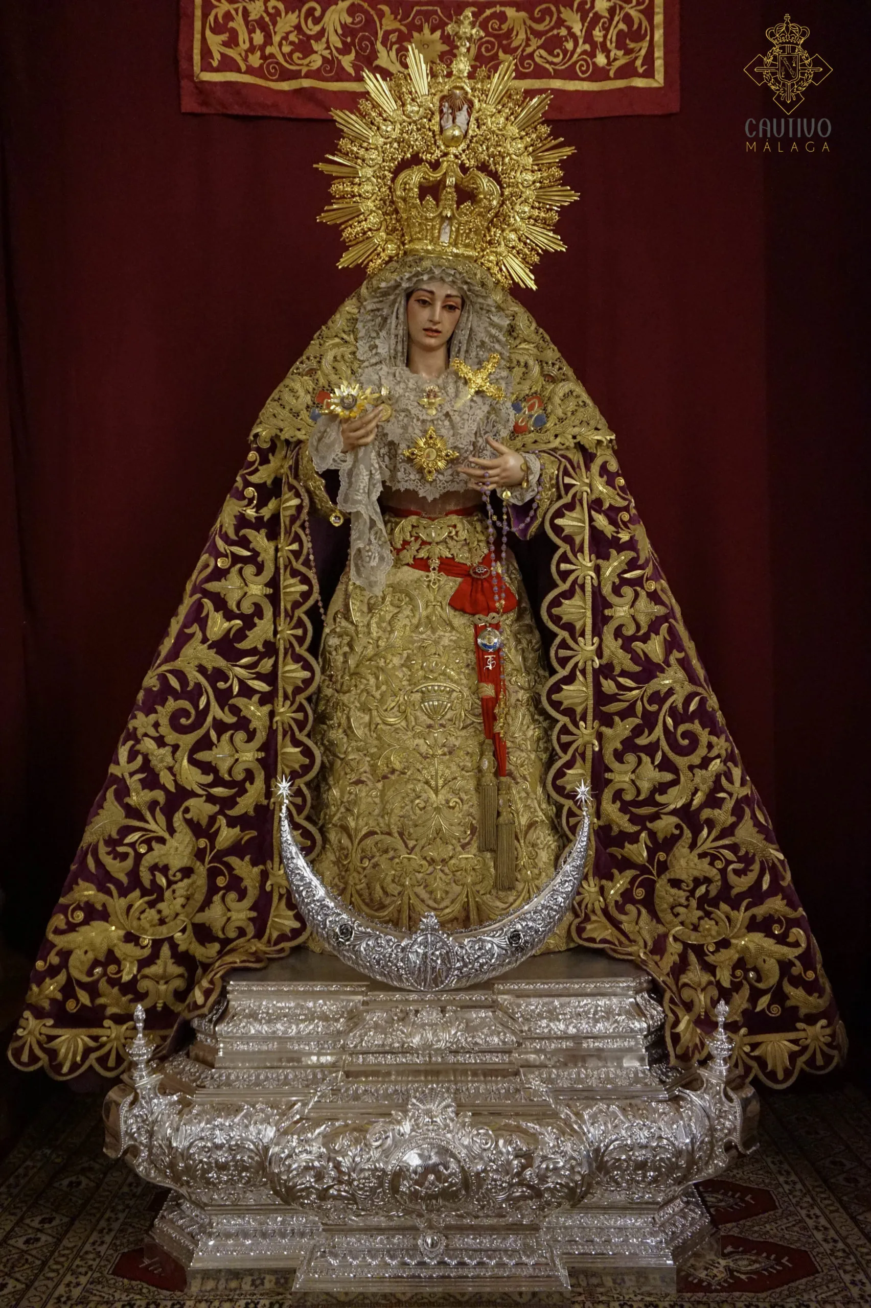 Orlada estatua religiosa de la Virgen María que lleva un manto burdeos bordado en oro y una corona dorada radiante, de pie sobre un elaborado pedestal de plata con una luna creciente.