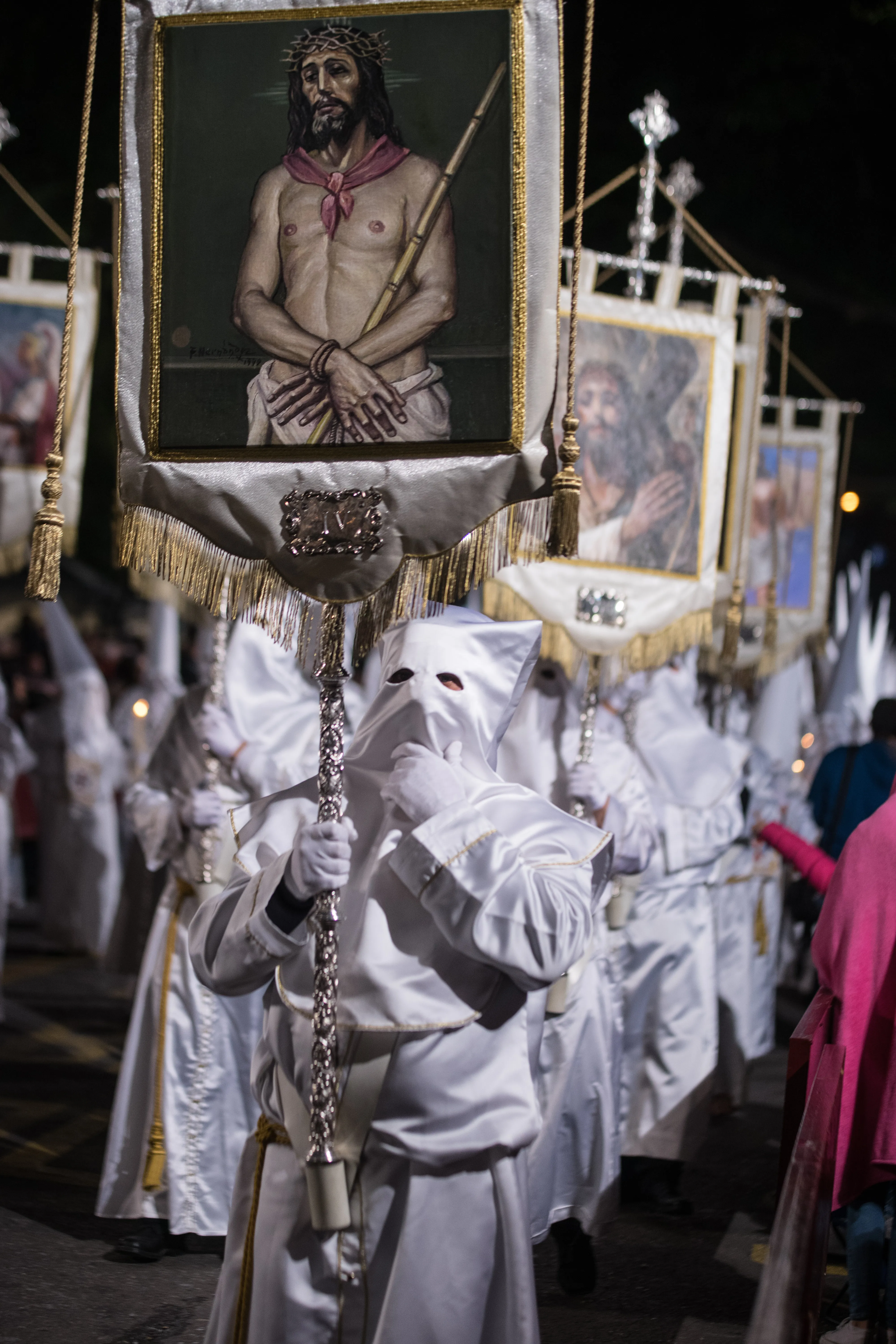 Procesión religiosa nocturna con penitentes vestidos con túnicas blancas y capirotes, portando grandes estandartes bordados que representan a Jesucristo con la corona de espinas.