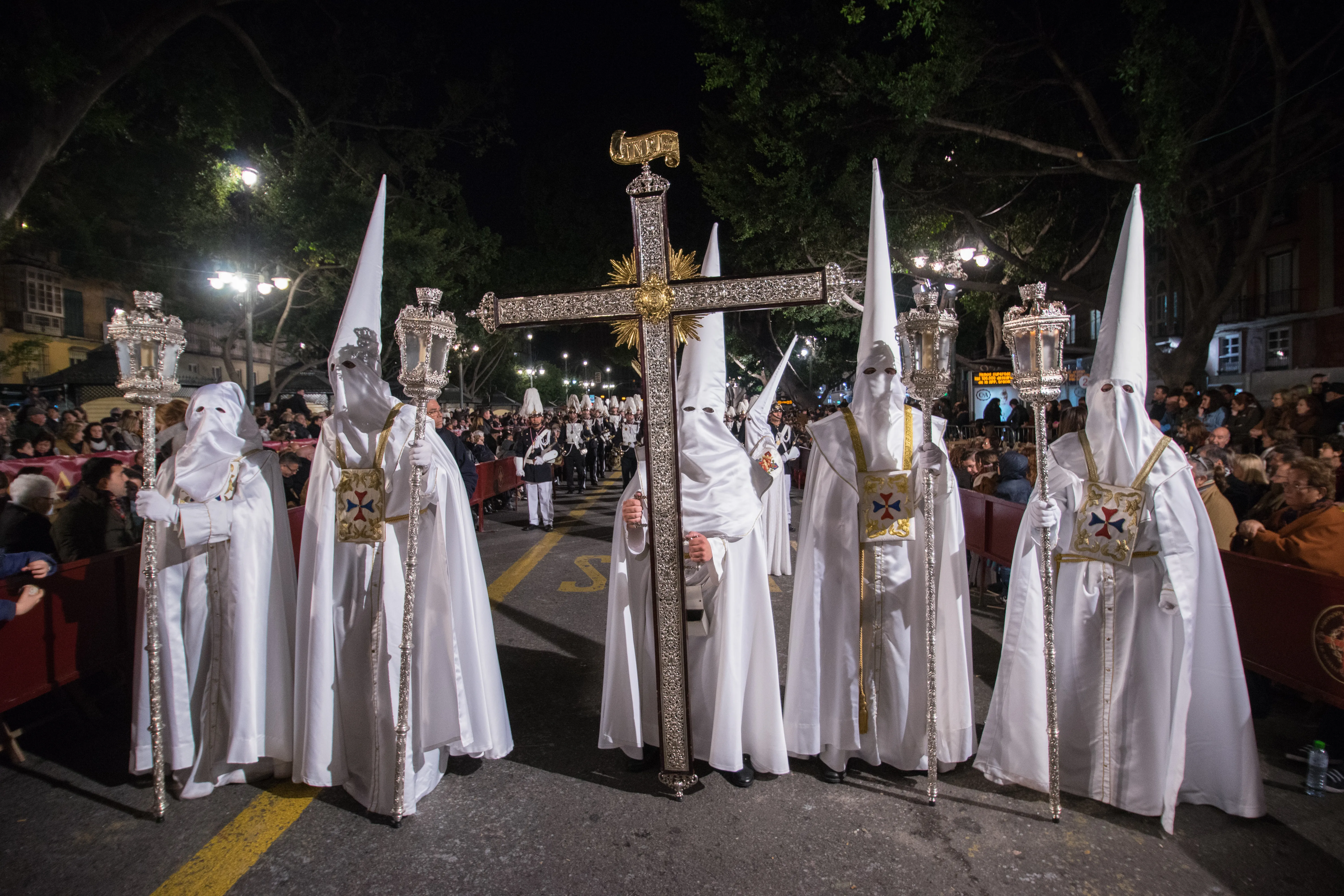 Group of nazarenos in white robes and tall pointed hoods carrying a large ornate processional cross and lanterns during a night procession in Málaga