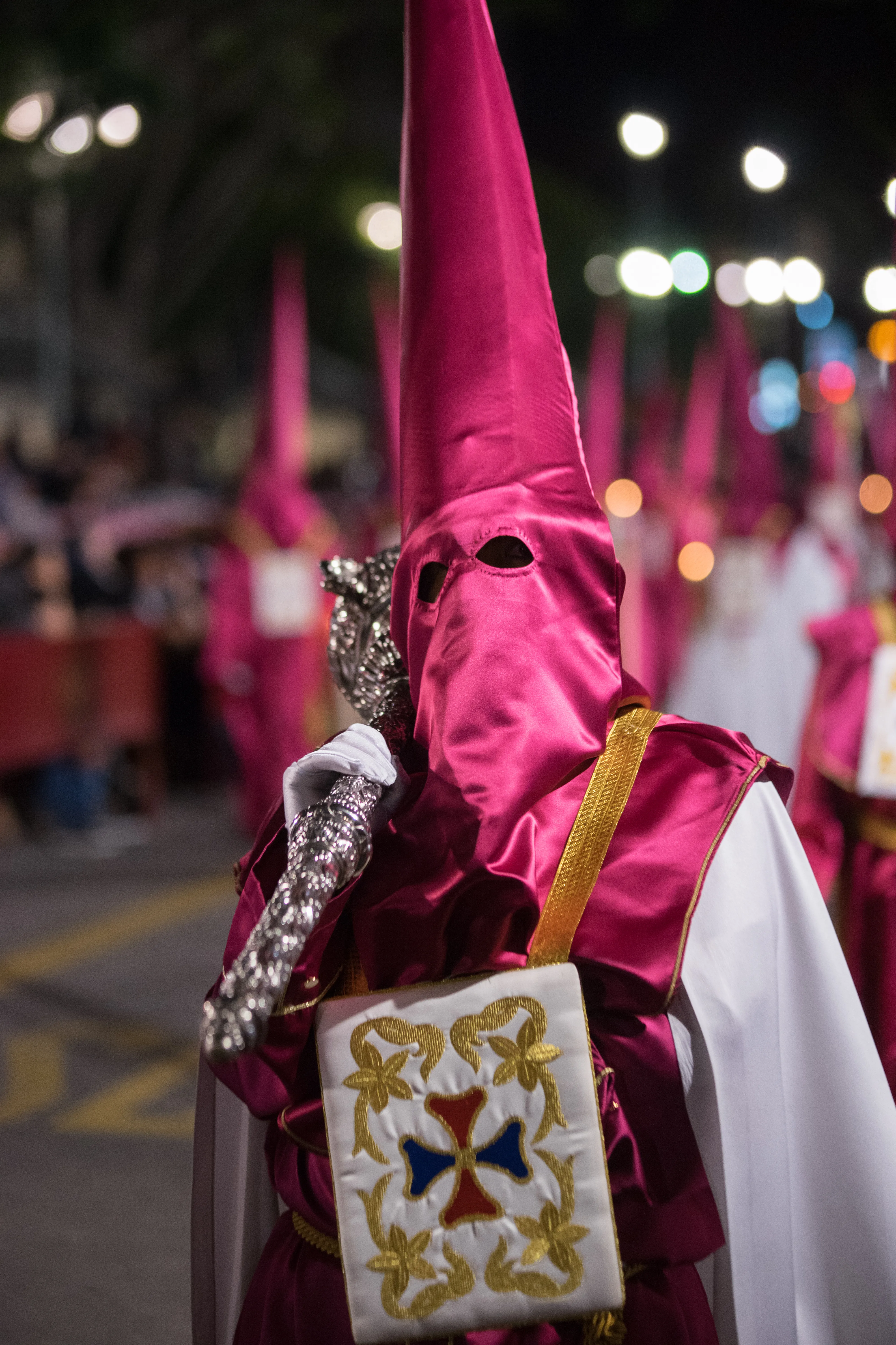 A nazareno in a tall magenta capirote hood and robe holds a silver staff and wears an embroidered white pouch with a cross during a nighttime Holy Week procession.