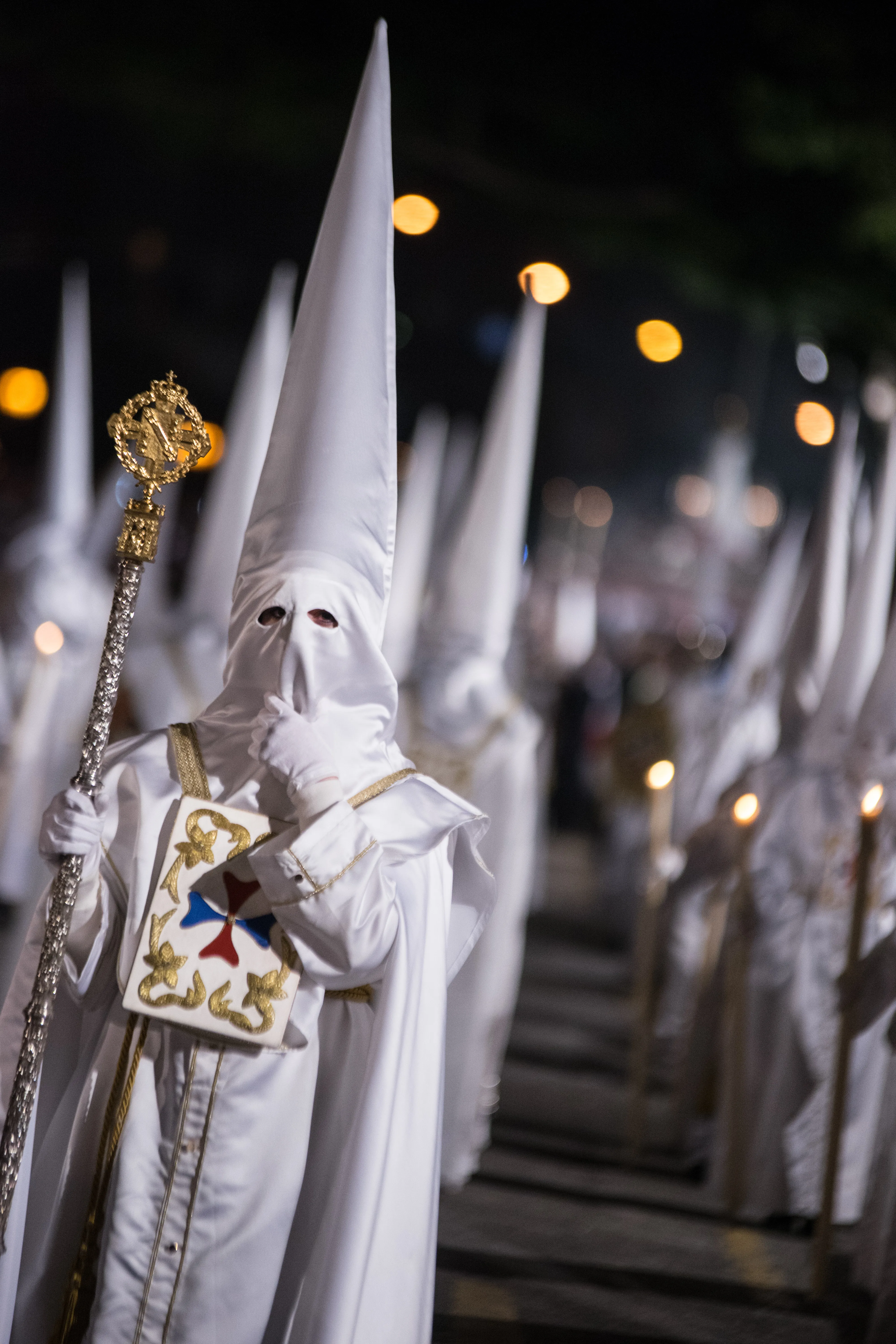 Nazareno con túnica y capirote blancos sosteniendo un báculo dorado durante una procesión nocturna de Semana Santa, con otras figuras encapuchadas y luces bokeh al fondo