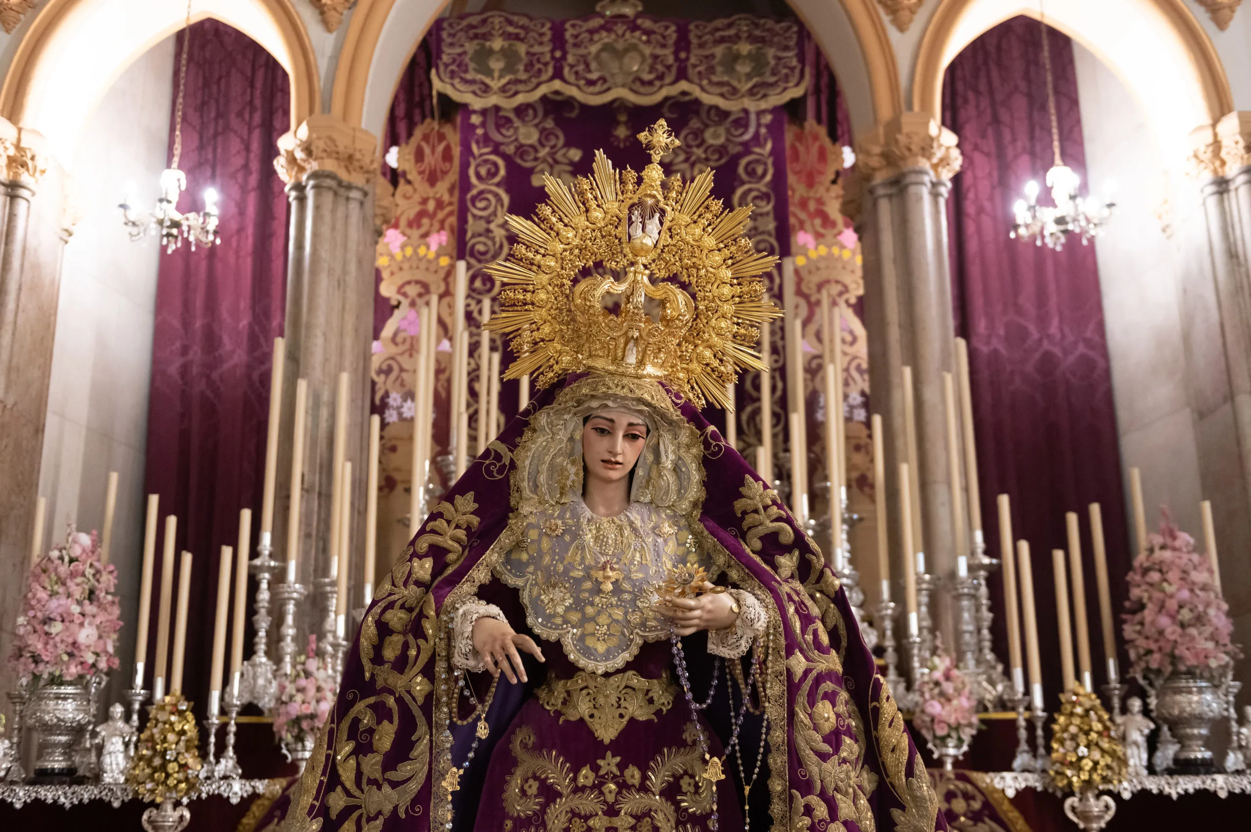 Una estatua devocional de la Virgen María con túnicas bordadas en oro y púrpura adornadas con un gran halo dorado, rodeada de velas blancas altas y flores rosas en el interior de una iglesia con columnas arqueadas.