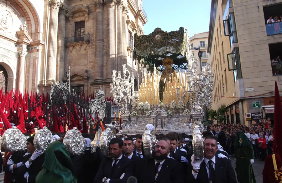 Un gran paso procesional de plata y oro que lleva un crucifijo, transportado por una calle frente a una catedral barroca, rodeado de costaleros con traje oscuro y nazarenos con túnicas rojas y capirotes plateados.