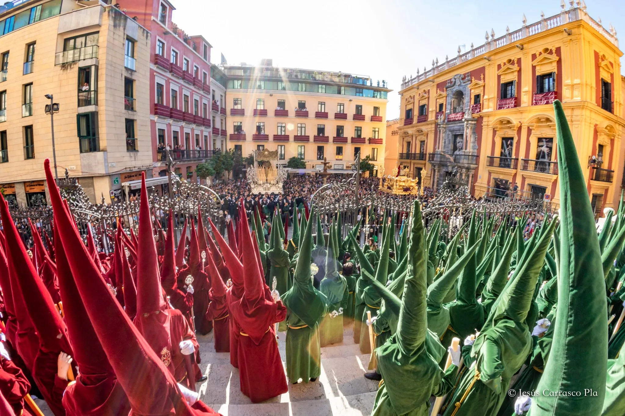 Procesión de Semana Santa con figuras encapuchadas con túnicas rojas y verdes que llenan una plaza rodeada de coloridos edificios históricos en Málaga