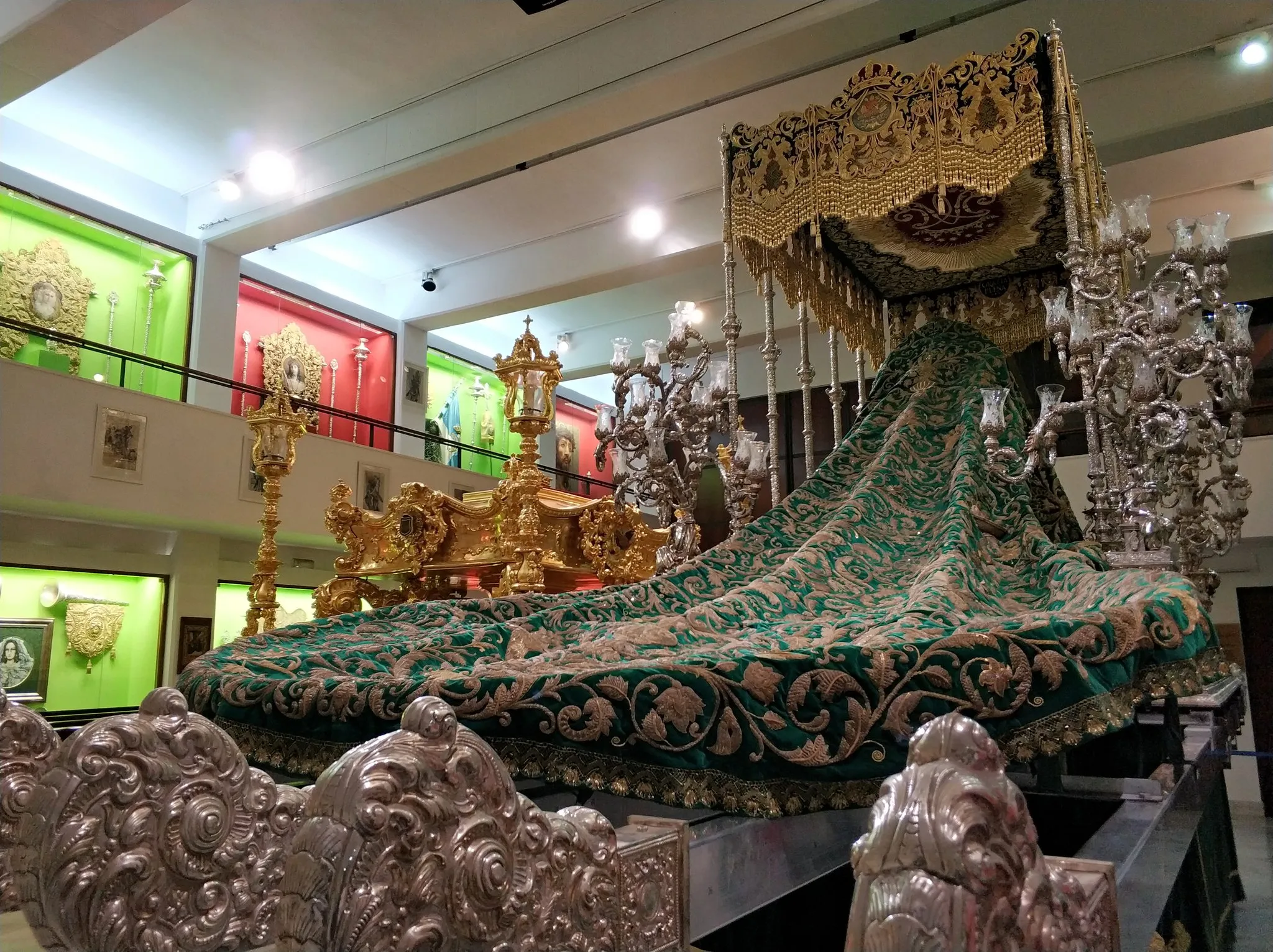 Ornado trono procesional de Semana Santa con un dosel de terciopelo verde ricamente bordado y candelabros de plata expuesto en una sala de exposiciones de un museo, con un trono dorado visible al fondo