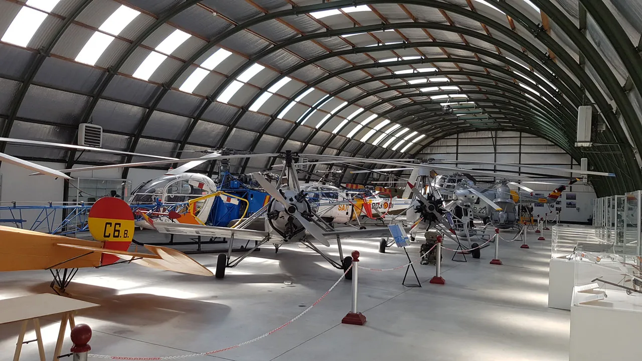 Interieur van een museumhangar met verschillende vliegtuigen tentoongesteld, waaronder helikopters en een klein vliegtuig, onder een gebogen metalen dak met dakramen.