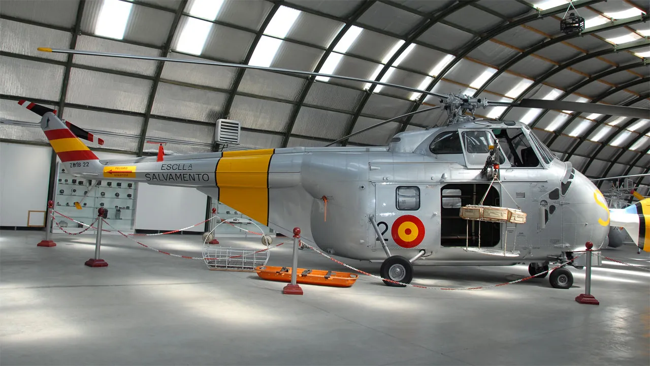 Een zilveren helikopter met gele en rode markeringen, tentoongesteld in een grote hangar