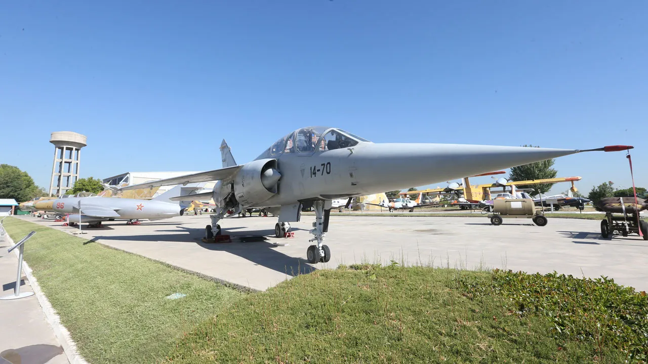 Een grijze militaire straaljager met het nummer 70 op de zijkant, geparkeerd op een betonnen ondergrond naast gras, met andere vliegtuigen en een toren op de achtergrond onder een blauwe hemel.