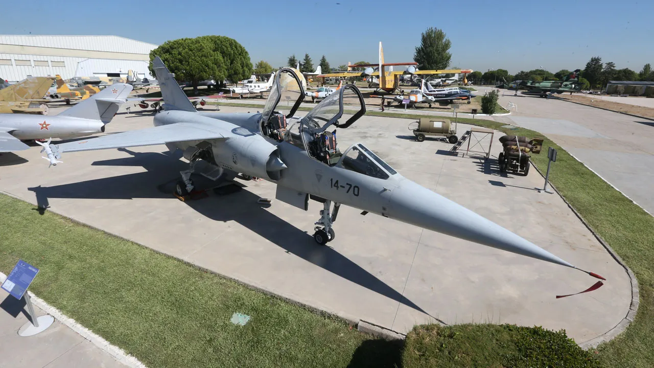 Een grijze militaire straaljager met open cockpit en zichtbare markeringen wordt buiten tentoongesteld op een betonnen ondergrond omringd door gras, met andere vliegtuigen en gebouwen op de achtergrond.