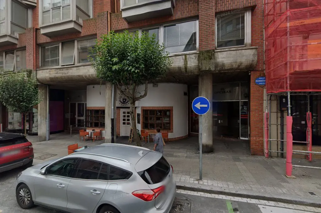 Exterior a nivel de calle de un edificio de ladrillo con voladizo de hormigón, un árbol, una señal de tráfico azul con flecha a la izquierda y un coche plateado aparcado delante