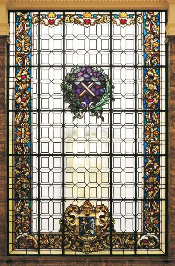 Glas-in-loodraam met een centraal embleem van gekruiste pikhouwelen en een krans, omgeven door sierlijke bloemmotieven en geometrische ontwerpen