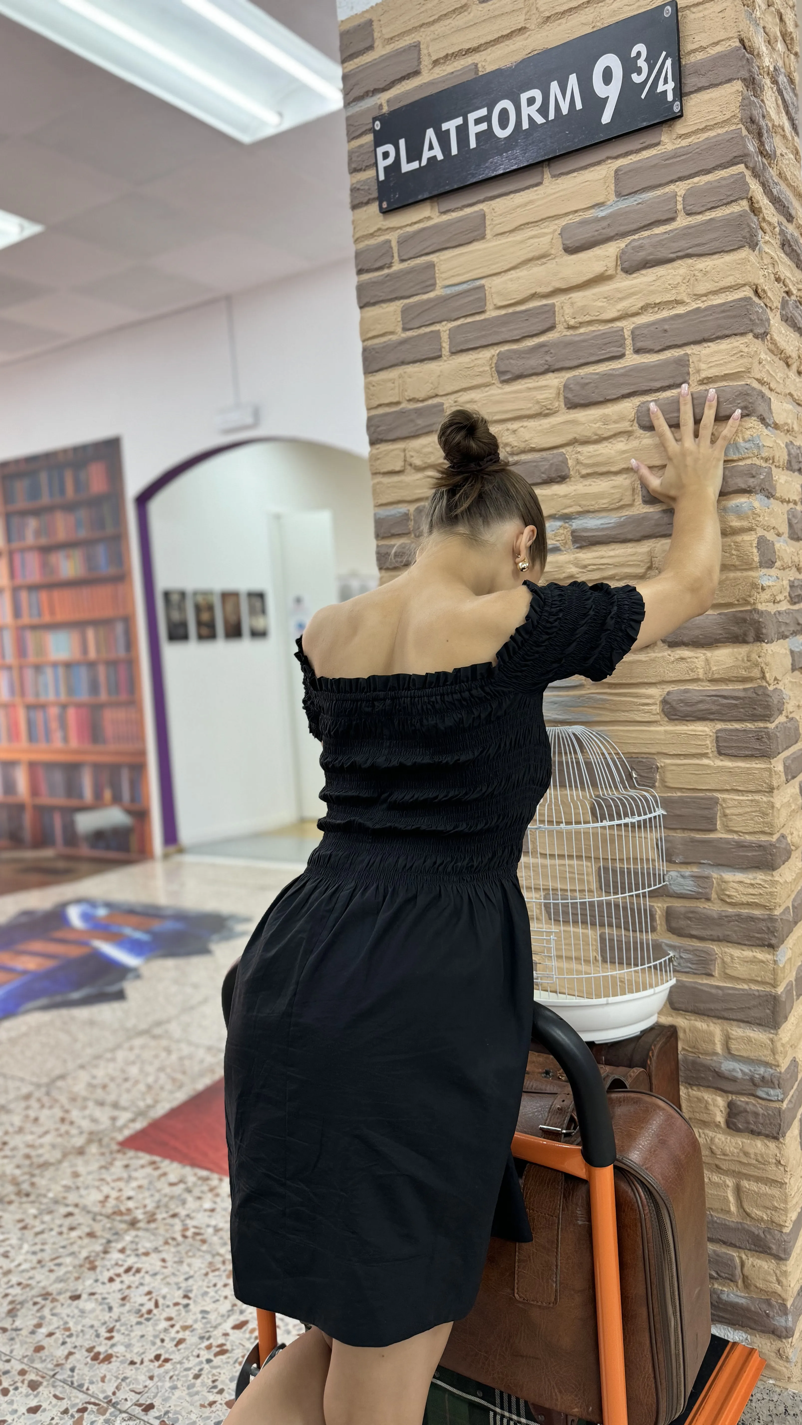 Una mujer con un vestido negro se apoya contra una pared de ladrillo falso bajo un cartel de Platform 9¾, empujando un carrito de equipaje con una maleta y una jaula de pájaros blanca.
