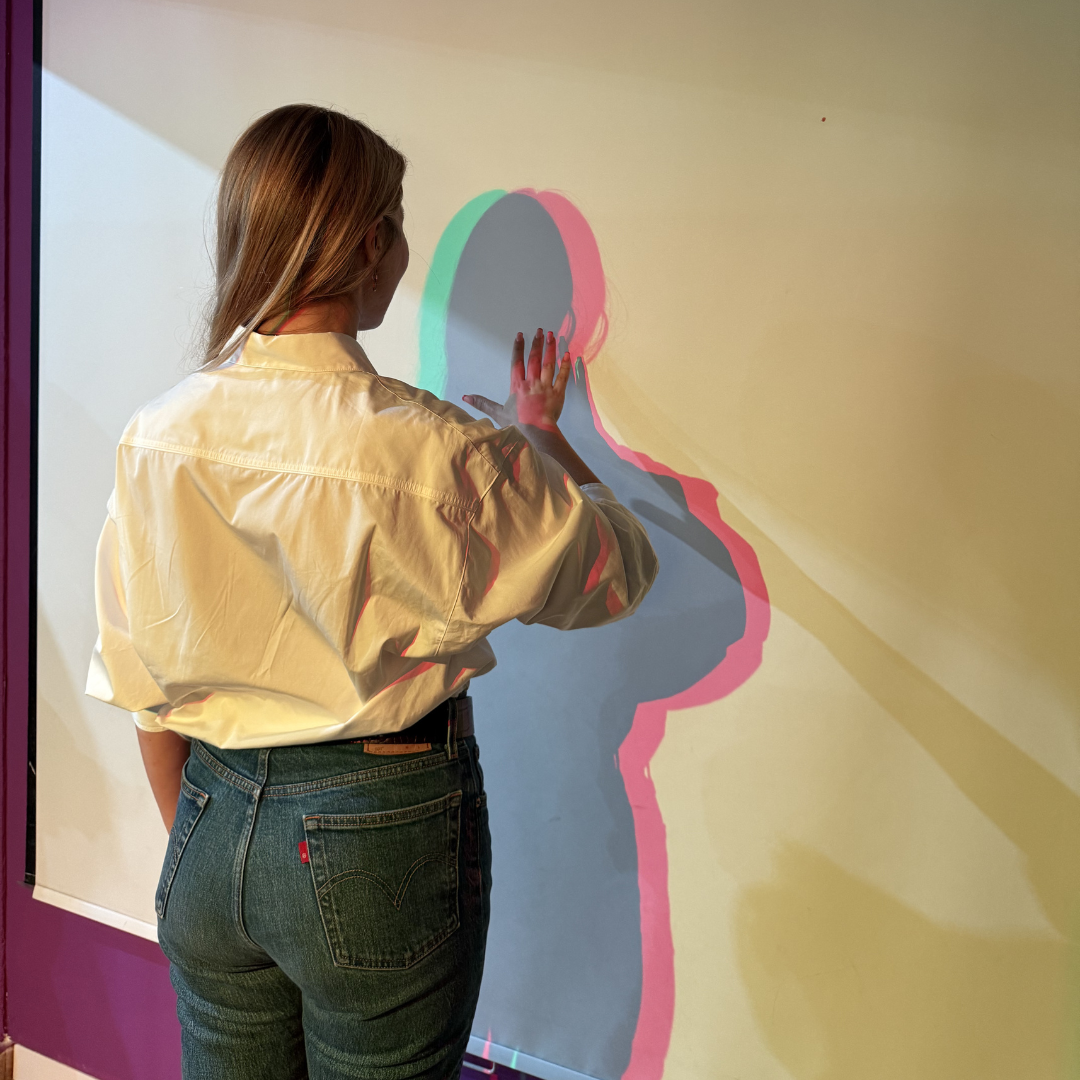 Una mujer está de pie frente a una pared blanca, proyectando una sombra multicolor en cian, magenta y azul gracias a fuentes de luz de colores.