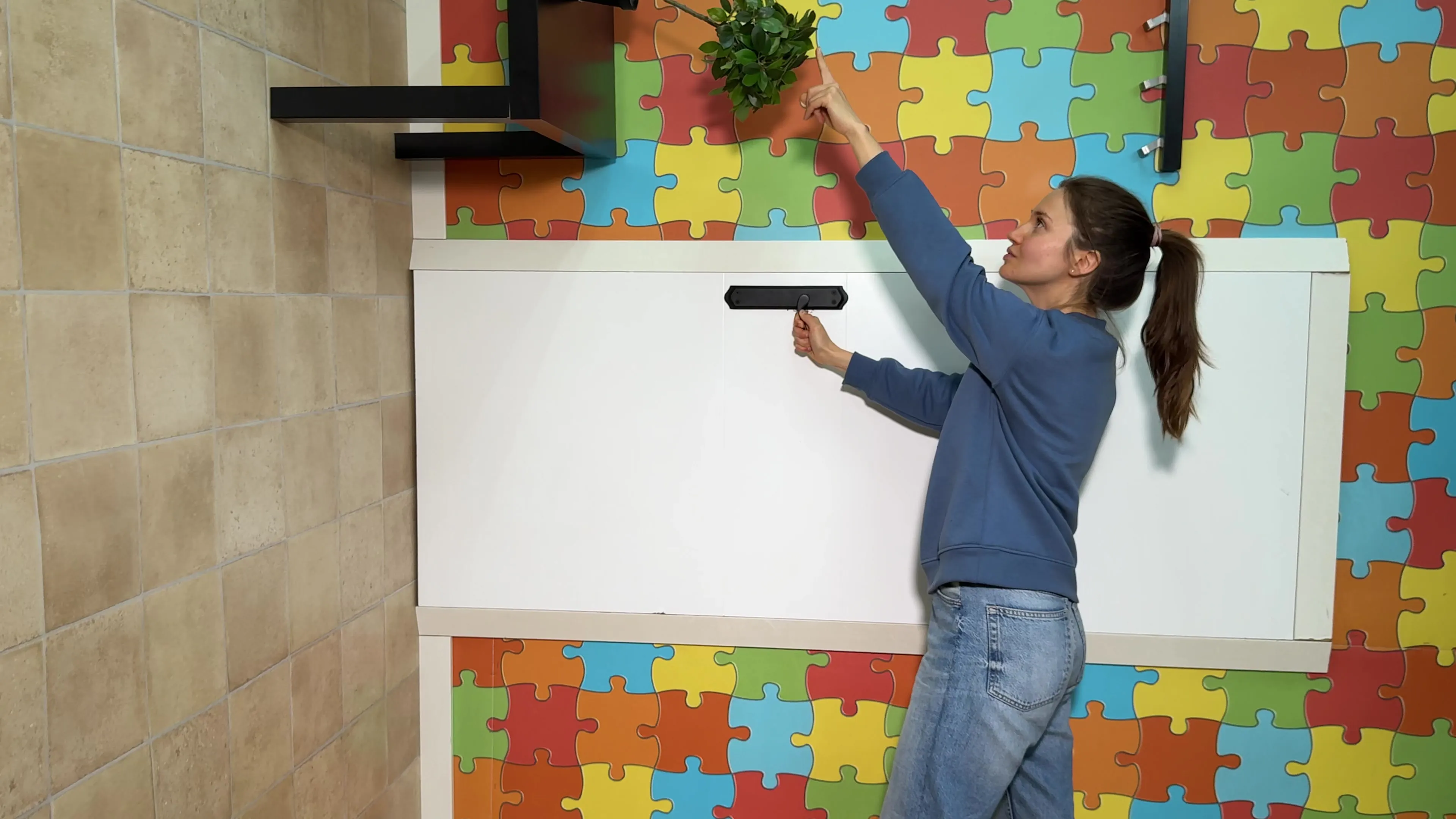 Una mujer alarga el brazo hacia arriba para regar una planta en un estante de pared dentro de una habitación con un colorido papel pintado de rompecabezas, creando una ilusión óptica que desafía a la gravedad en el Museo de la Imaginación.