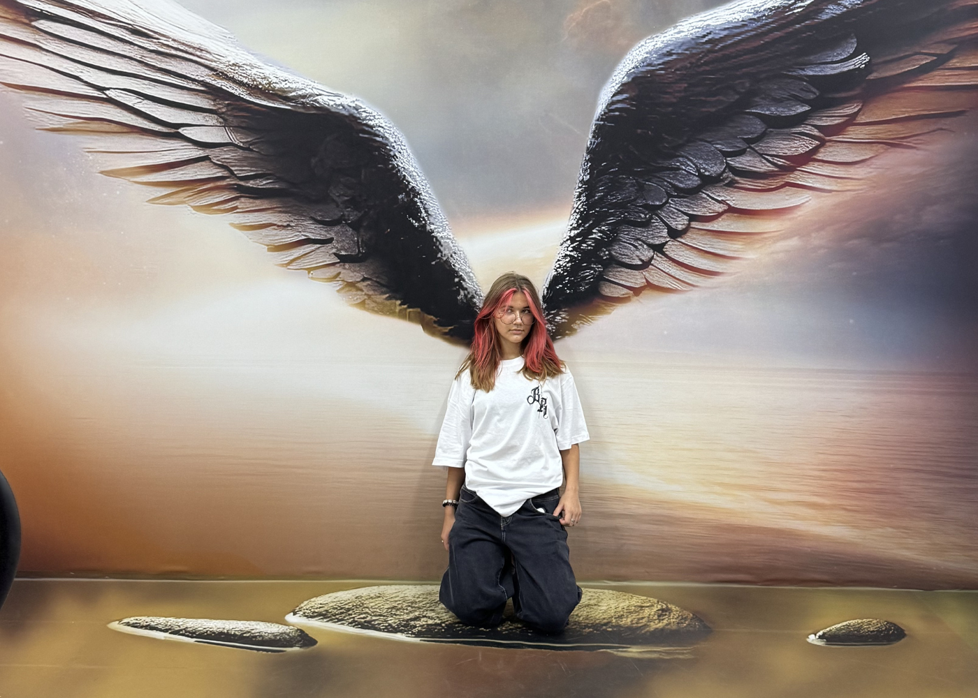 Una persona arrodillada frente a un gran mural de alas de ángel, posicionada para crear la ilusión óptica de tener alas.