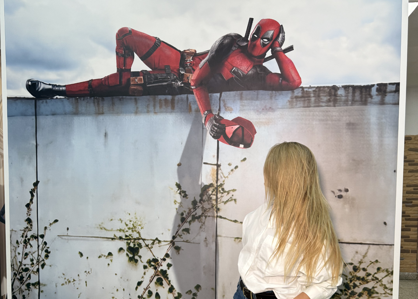 Una mujer de cabello largo y rubio está de pie frente a un gran mural que representa al personaje de cómic Deadpool recostado sobre un muro de hormigón.