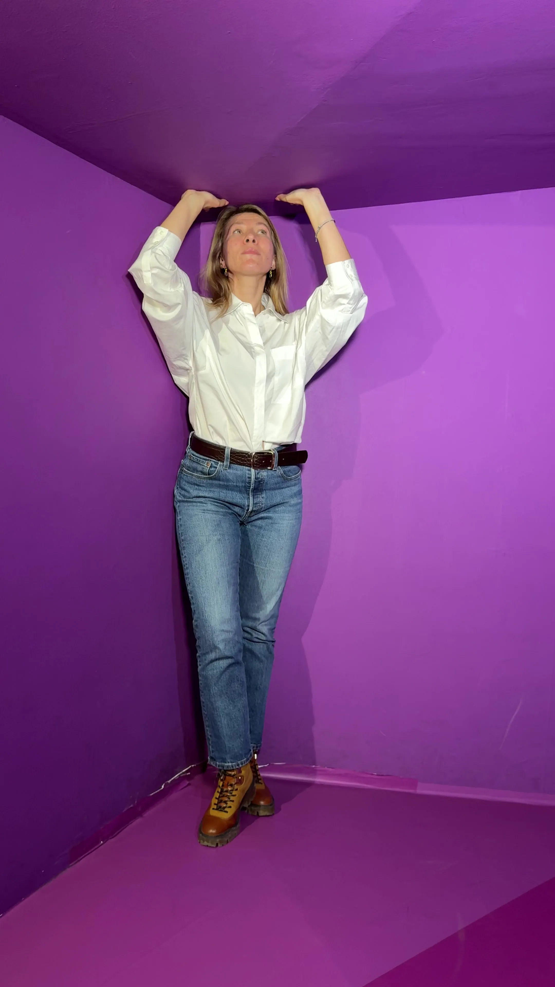 Una mujer con camisa blanca y pantalones vaqueros azules está de pie en la esquina de una habitación morada, estirándose para tocar el techo con ambas manos en una exposición de ilusión óptica.
