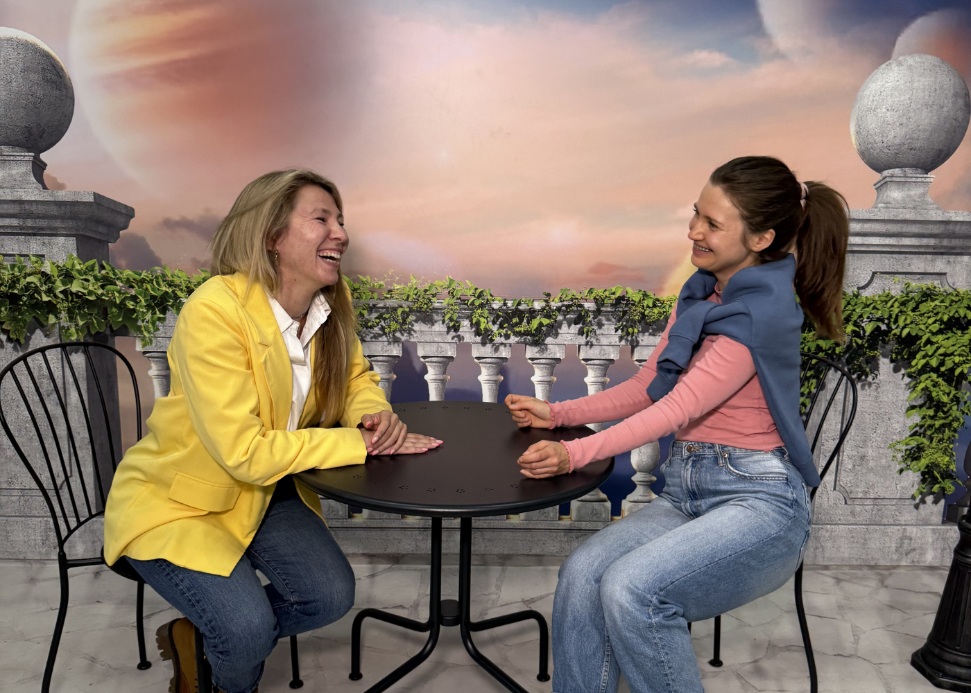 Two women sitting at a small round table in a forced perspective room with a painted terrace backdrop featuring balustrades, decorative spheres, and a sky, creating an optical illusion of different sizes.