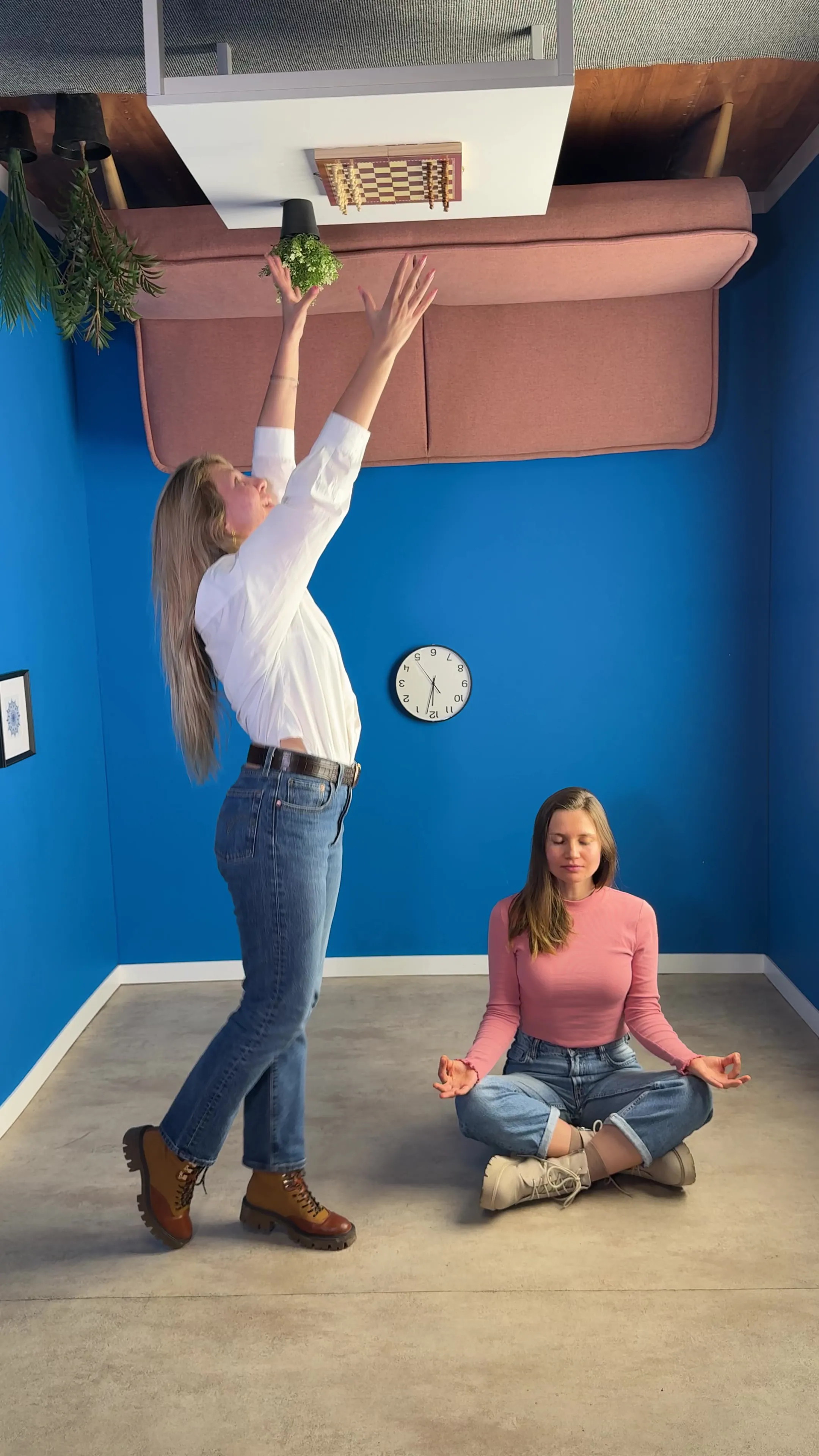 Dos mujeres posan en una habitación de ilusión óptica girada con paredes azules, una estirándose hacia los muebles montados en el techo mientras la otra está sentada en posición de loto meditando en el suelo.