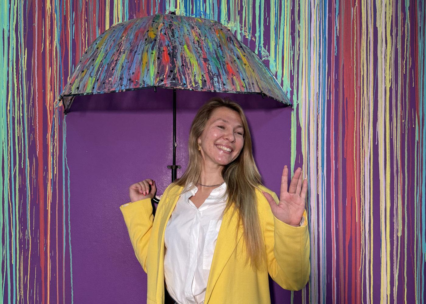 A smiling woman in a yellow jacket holds a colorful paint-splattered umbrella in front of a purple wall with multicolored vertical paint drip patterns.
