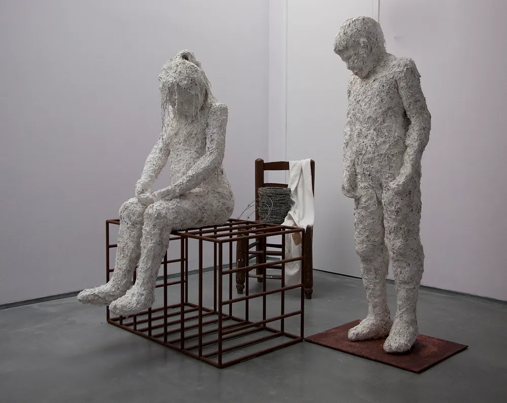 Dos esculturas de figuras humanas blancas con textura en una galería, una sentada sobre un marco de rejilla metálica y otra de pie sobre una base, con una silla de madera al fondo.