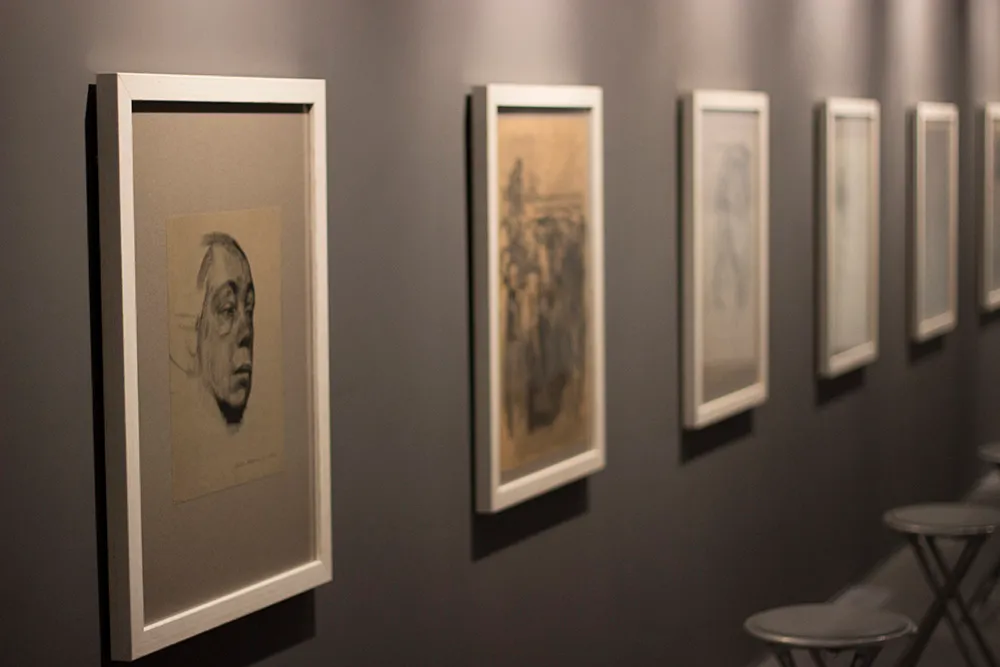 Obras de arte enmarcadas colgadas en fila en una pared gris oscuro de un museo, incluyendo un grabado de un retrato y otros dibujos, con taburetes visibles en primer plano