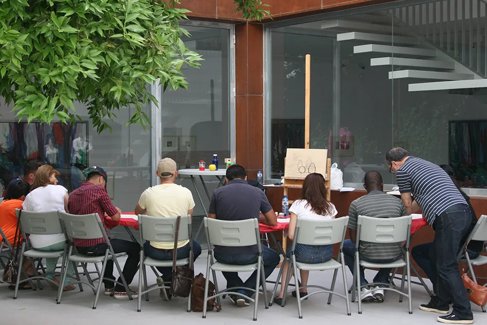 Grupo de personas sentadas en mesas al aire libre participando en un taller de arte con un caballete y un lienzo visibles, frente a un edificio moderno de cristal con una escalera