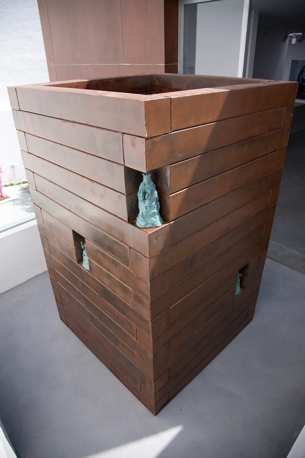 Gran escultura de metal en forma de caja color óxido hecha de placas horizontales con pequeñas aberturas empotradas que contienen figurillas de color azul verdoso, expuesta en una galería de museo.