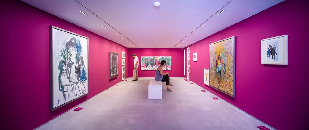 Vista panorámica amplia de una sala de museo con paredes de color magenta brillante, que presenta grandes pinturas expresionistas enmarcadas y un visitante sentado en un banco blanco en el centro.