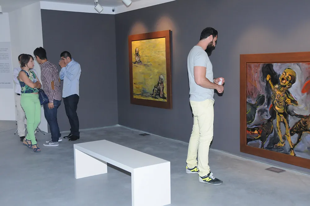 Visitantes de pie observando grandes pinturas expresionistas enmarcadas en paredes grises dentro de una sala de museo con un banco blanco
