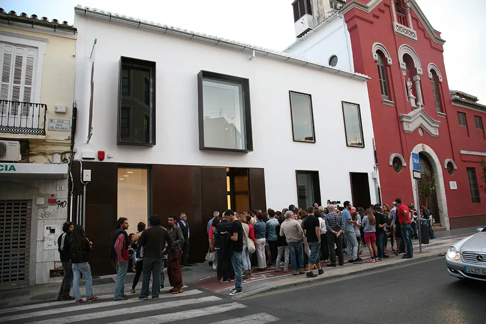 Edificio de museo blanco de dos pisos con grandes ventanales de marco oscuro y una gran multitud de personas paradas en la acera frente a él, junto a una iglesia roja.