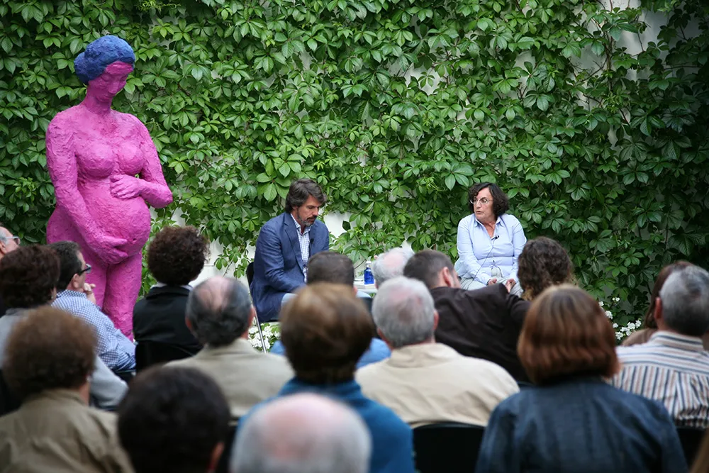 Una gran escultura rosa de una mujer embarazada con un pañuelo azul en la cabeza se encuentra junto a dos altavoces en un evento al aire libre, con el público sentado frente a una pared cubierta de hiedra.