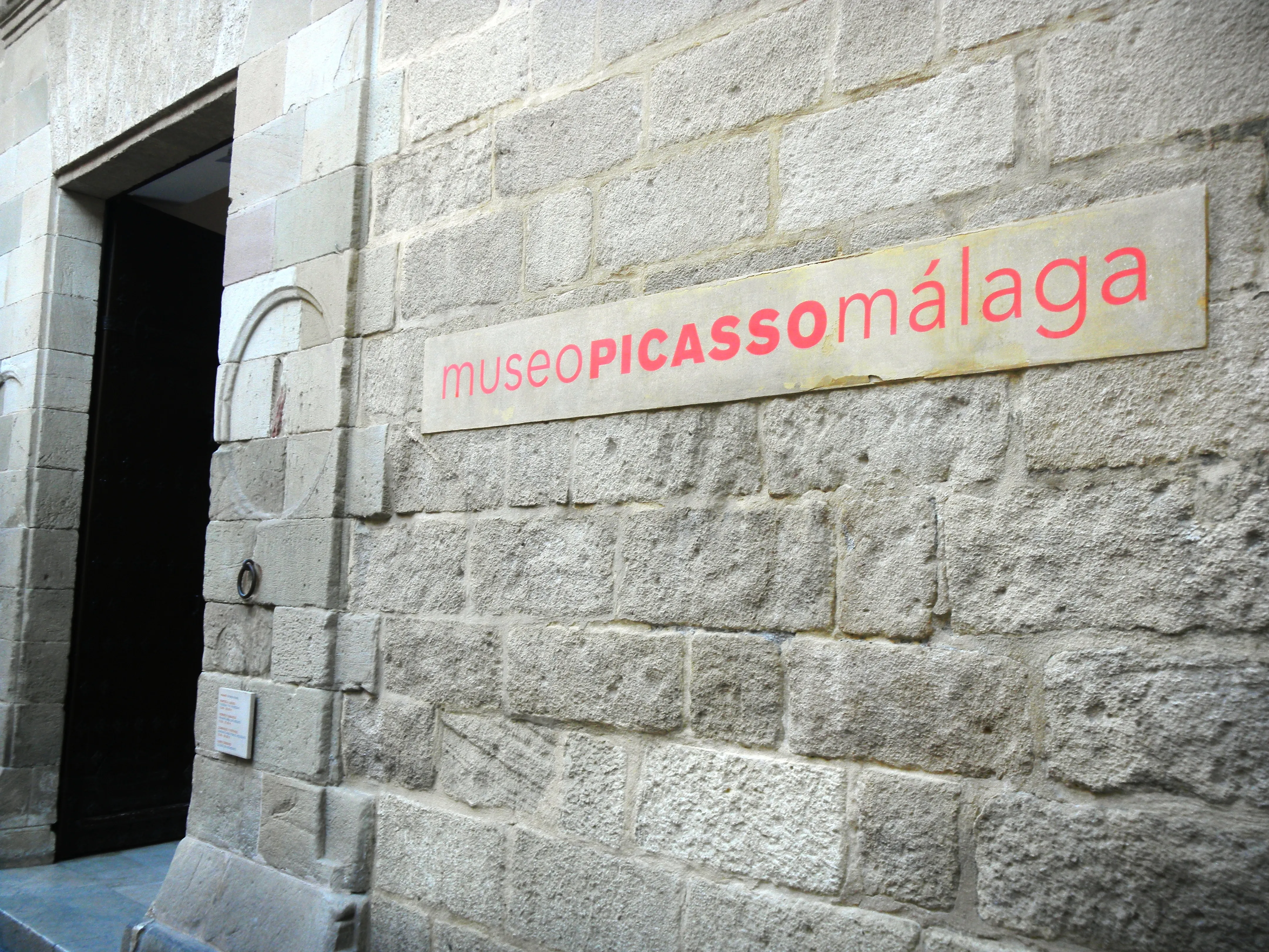 Muro de piedra del Museo Picasso Málaga con un letrero rojo y rosa que dice 'museoPICASSOmálaga' junto a una entrada oscura.