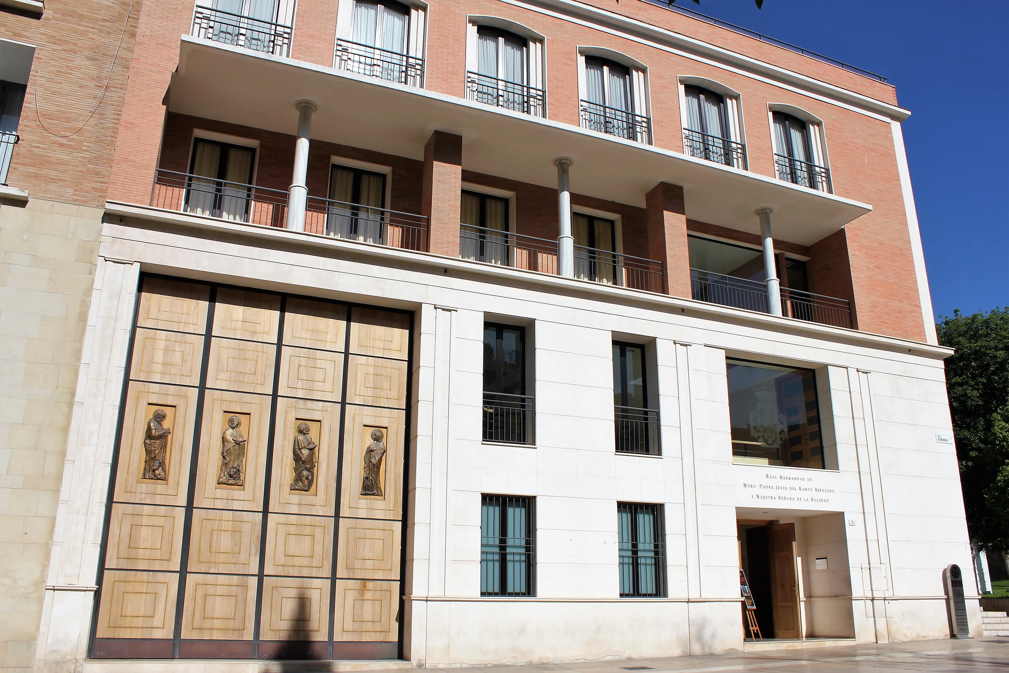 Fachada exterior de un edificio de museo que muestra una gran puerta de madera con cuatro paneles de figuras religiosas, pisos superiores de ladrillo y blanco con balcones y columnas bajo un cielo azul