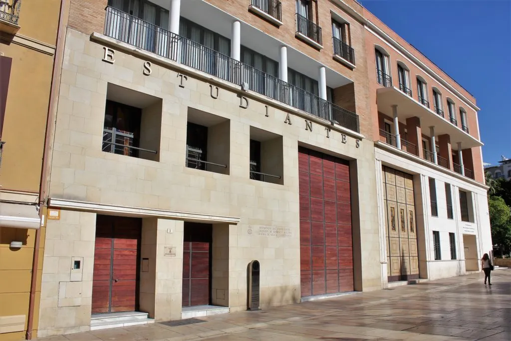 Fachada soleada de un gran edificio beige con la inscripción 'ESTUDIANTES' en el nivel superior, que cuenta con grandes puertas de madera de color marrón rojizo y balcones.