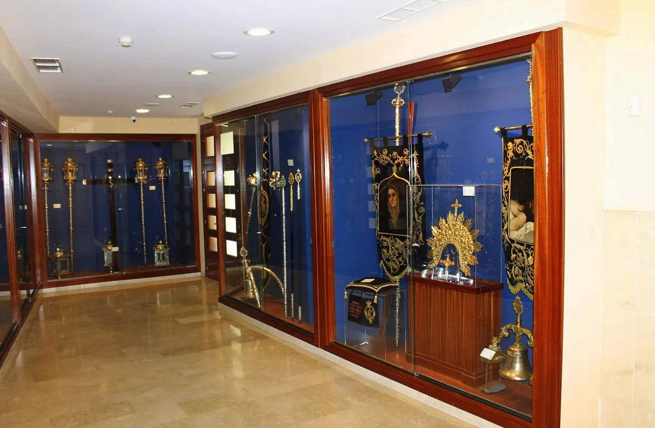 Interior de museo con vitrinas de cristal que contienen estandartes religiosos, báculos dorados y artefactos sobre fondos azules.