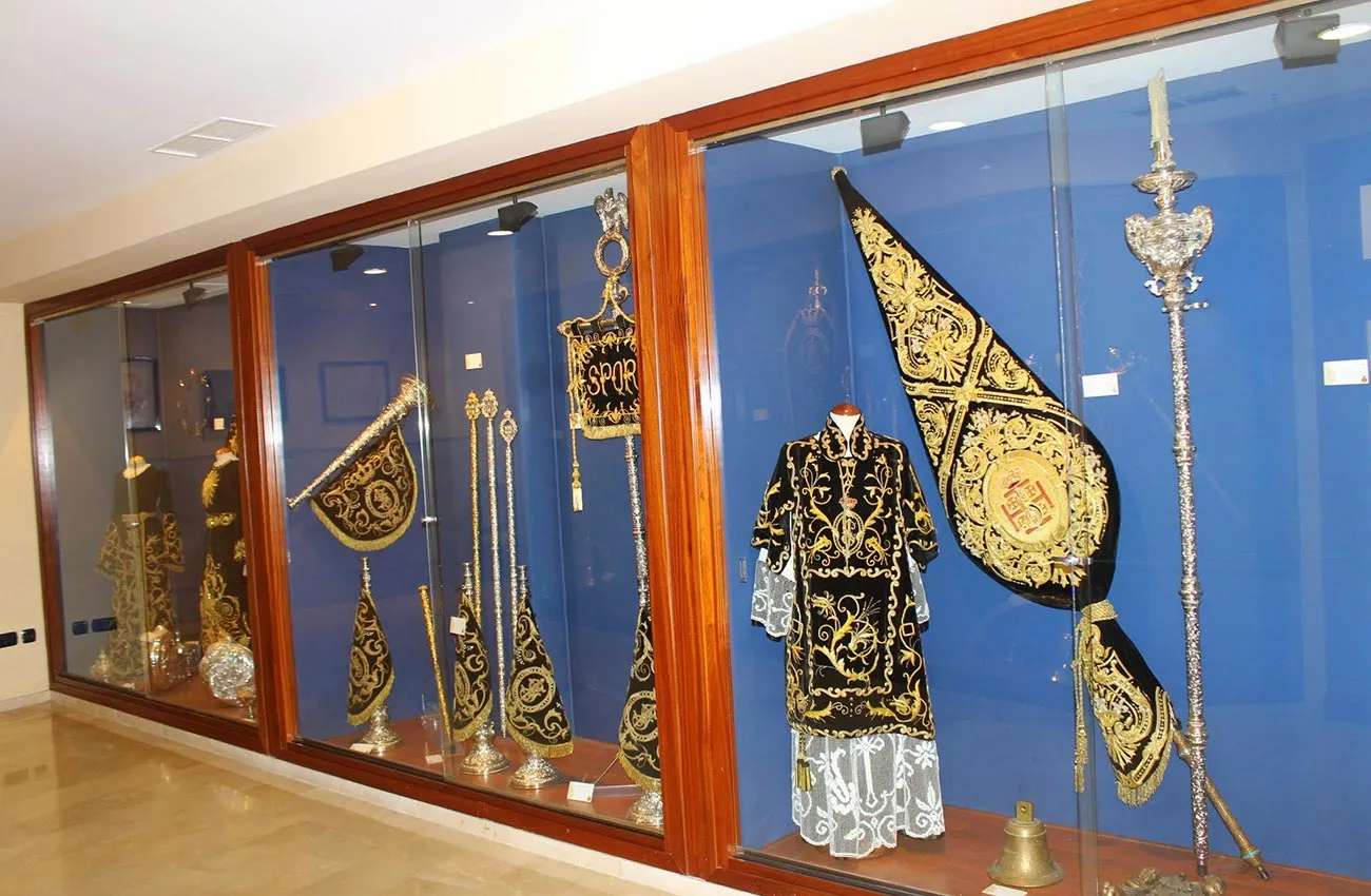Vitrina de cristal en un museo que exhibe ornamentados ornamentos de Semana Santa bordados en oro y negro, estandartes y bastones ceremoniales sobre fondos azules.