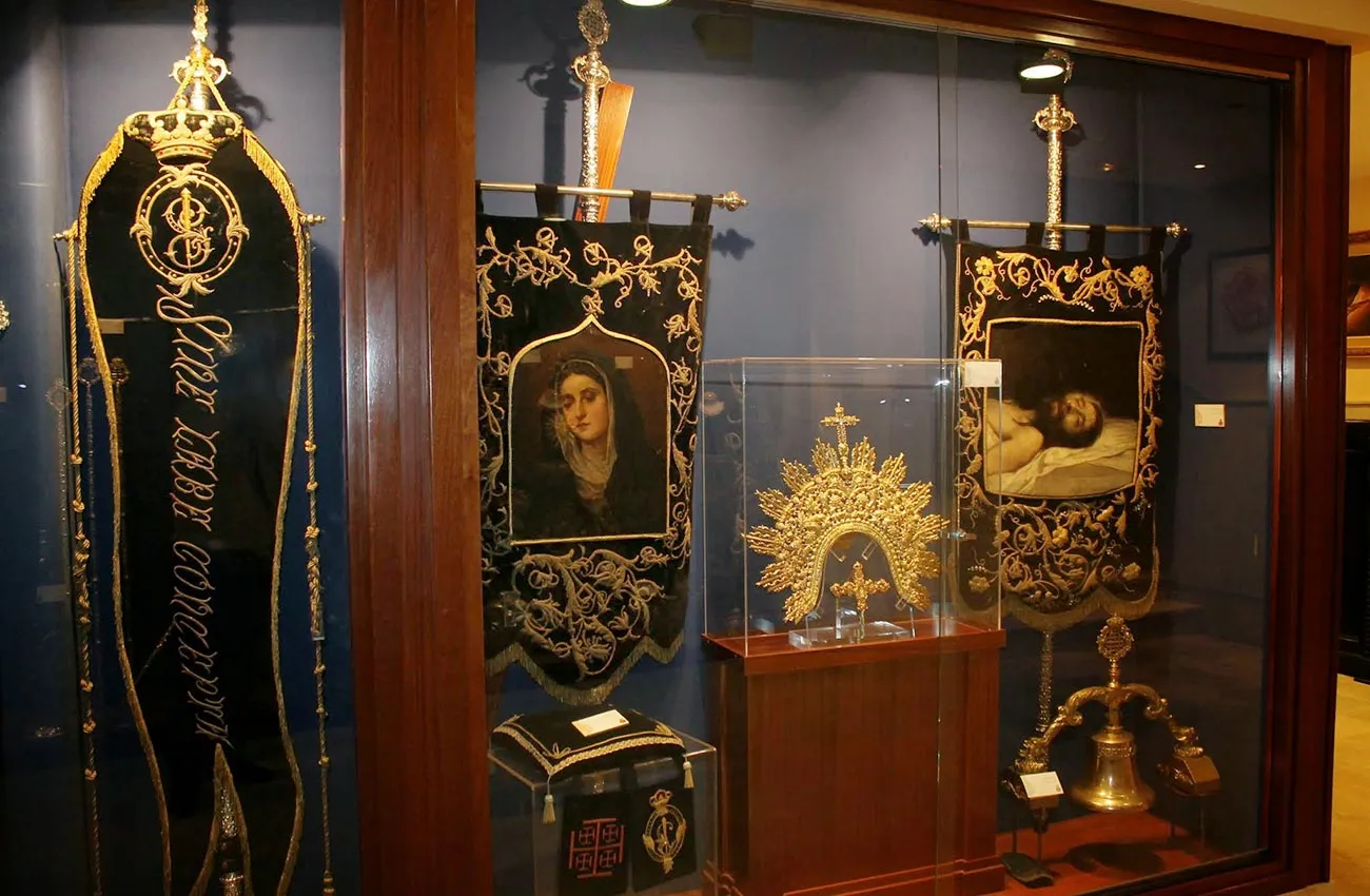 Vitrina de museo con estandartes procesionales altos bordados en negro, una corona radiante dorada en una caja de cristal y una campana de latón sobre un soporte de madera.