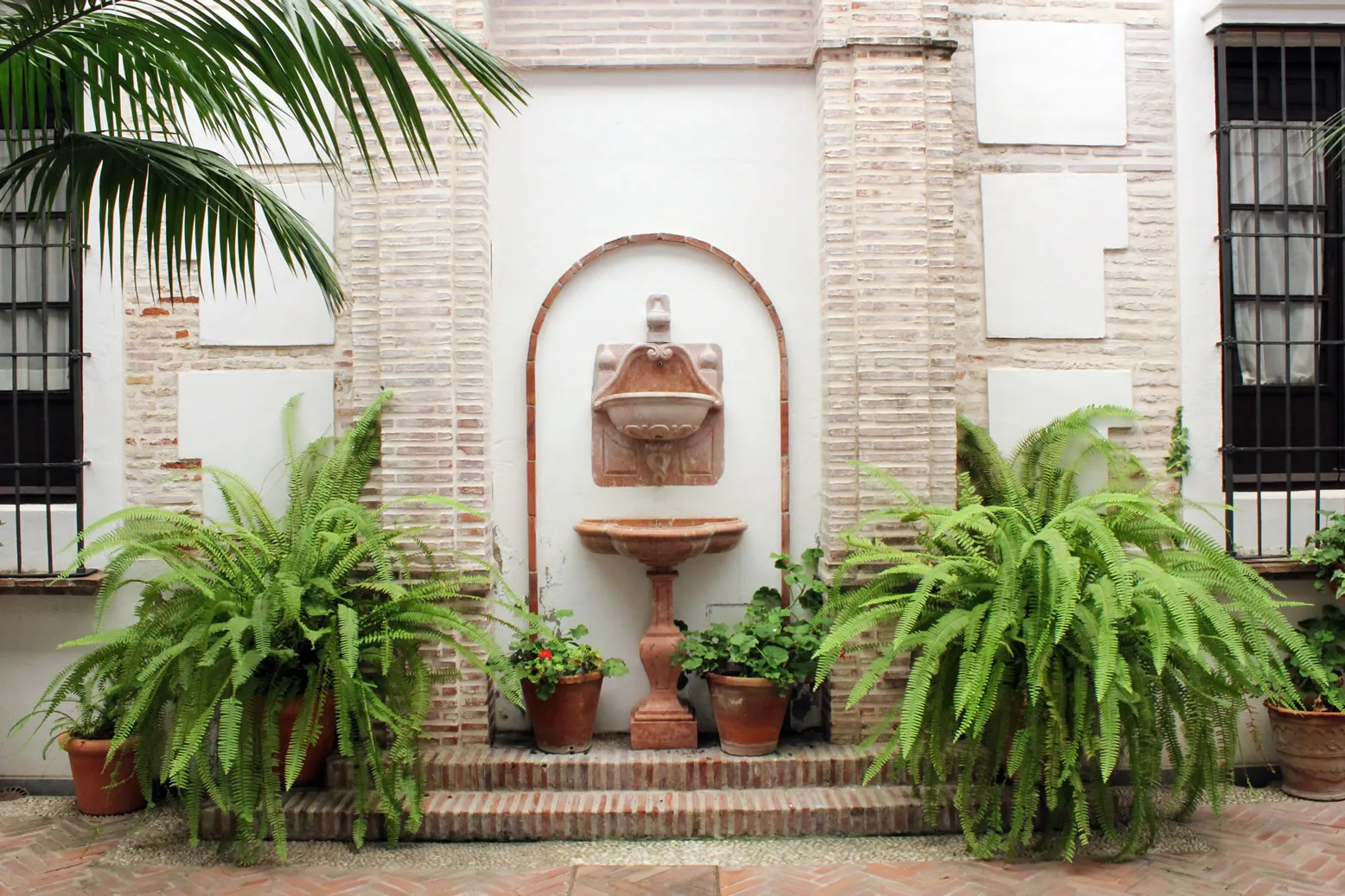 Un patio de ladrillo con una hornacina de fuente arqueada, helechos en macetas a ambos lados y ventanas con rejas de hierro en el Museo de la Semana Santa de Málaga.