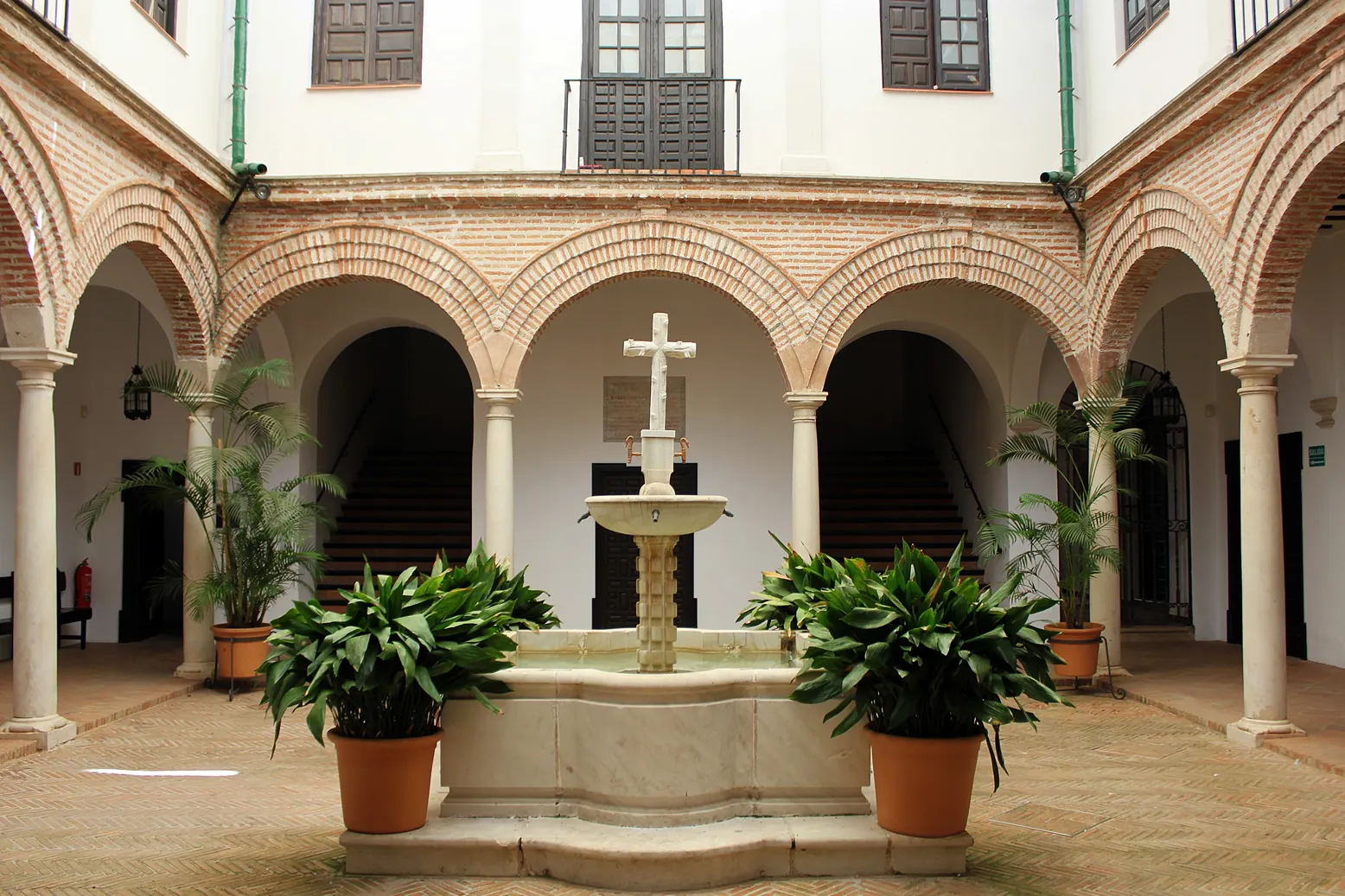Patio interior con una fuente central de piedra coronada por una cruz, rodeado de arcos de ladrillo, columnas blancas y grandes plantas en macetas.