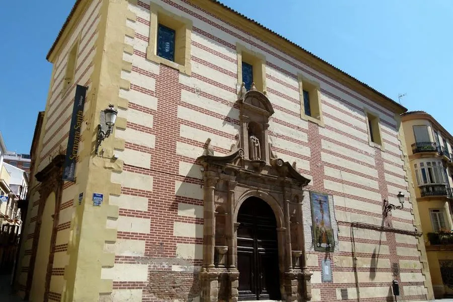 Fachada exterior del Museo de la Semana Santa de Málaga con muros de ladrillo rojo y bandas horizontales blancas, junto a una ornamentada portada barroca de piedra