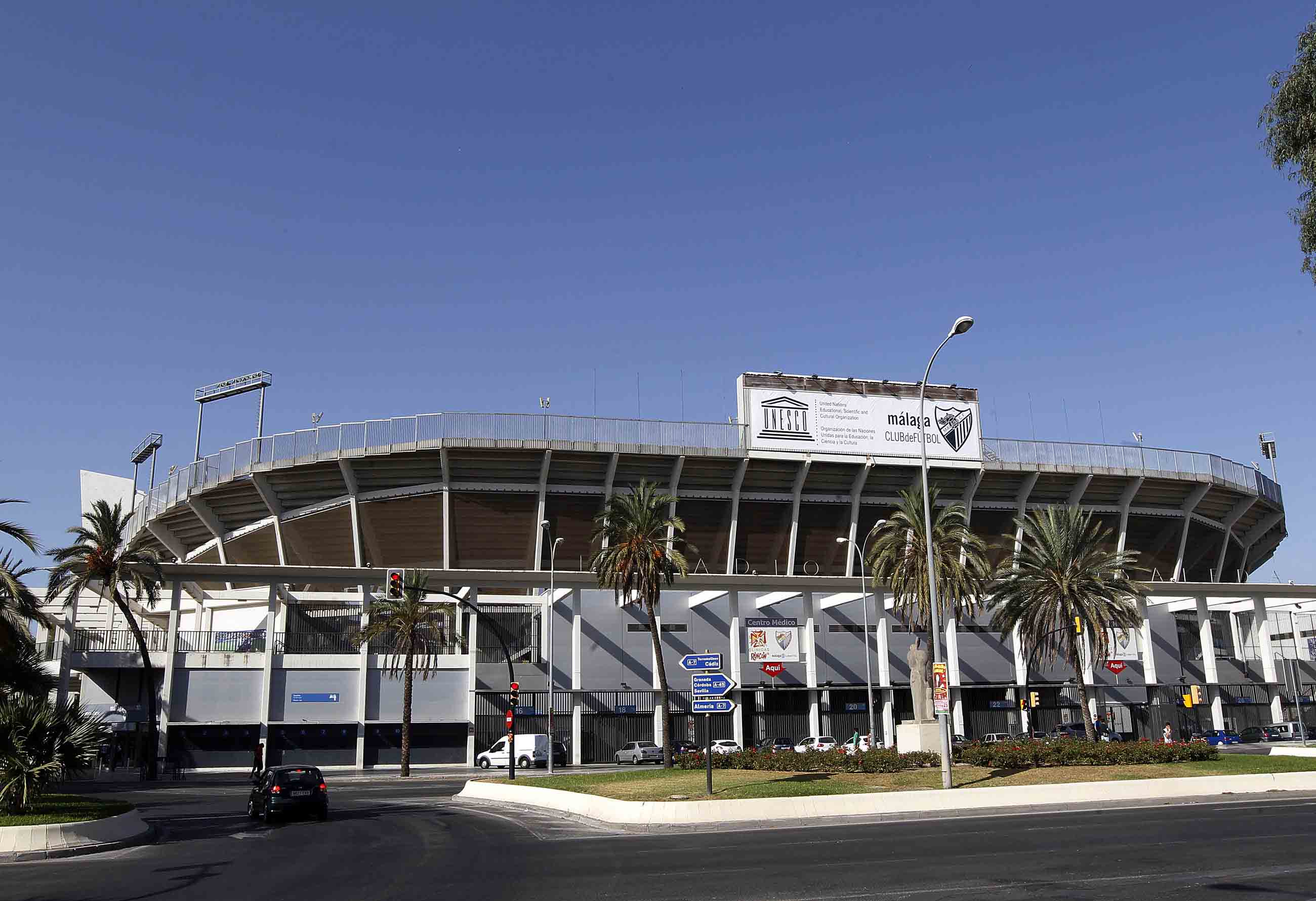Vista exterior del estadio La Rosaleda con una gran pancarta del Málaga CF en la fachada, palmeras y un cielo azul despejado