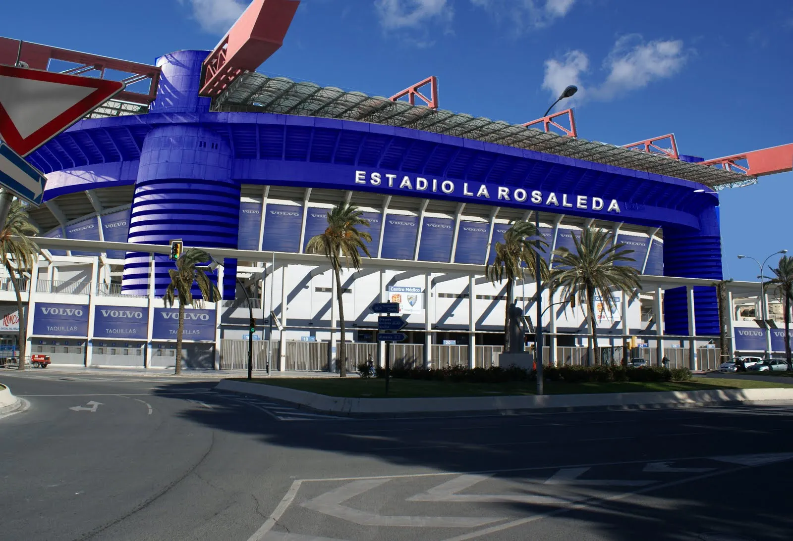Fachada exterior azul del Estadio La Rosaleda con letras blancas y palmeras al frente bajo un cielo azul