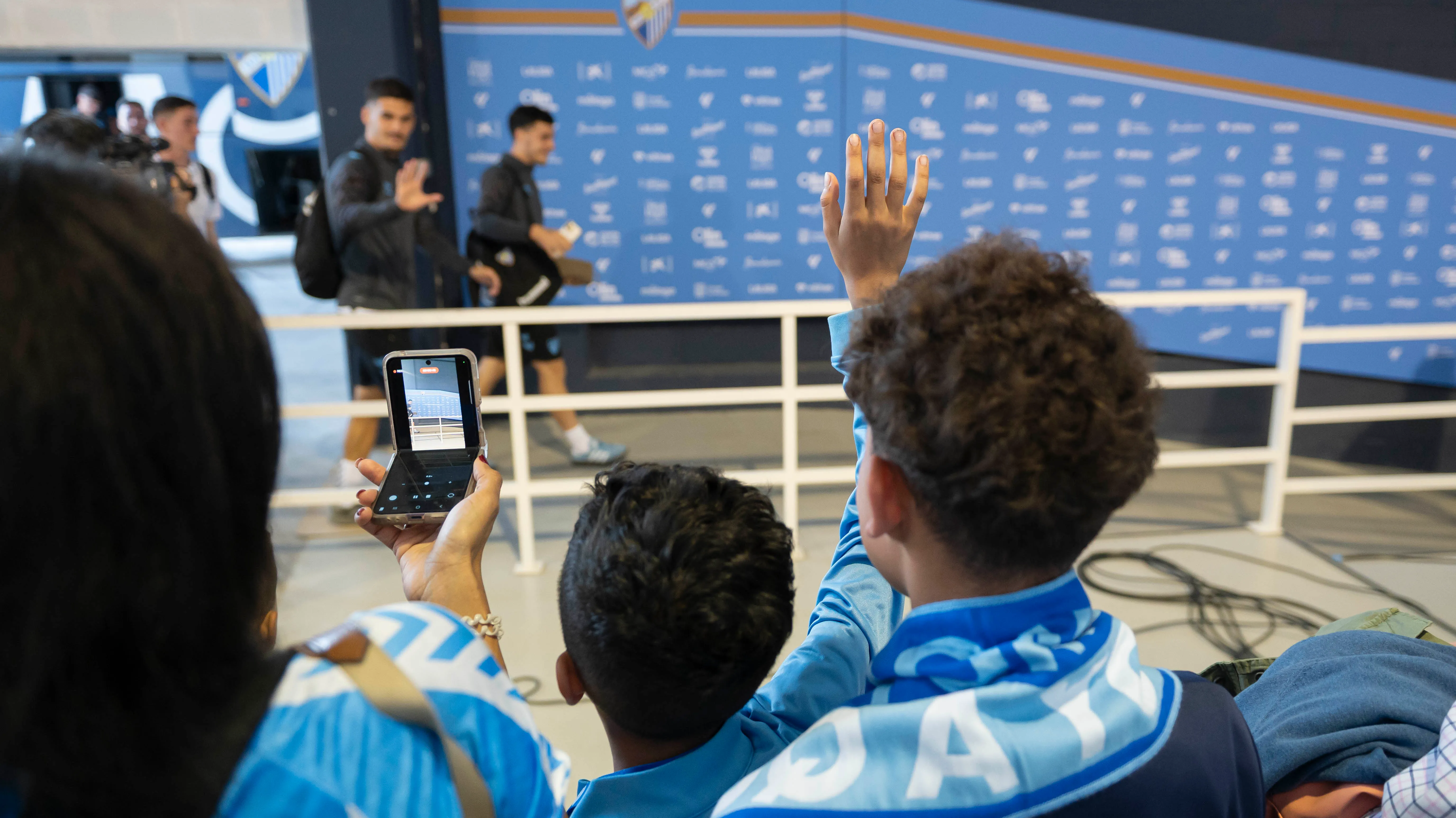 Aficionados con bufandas azules del Málaga CF saludan y levantan sus teléfonos para fotografiar a los jugadores que pasan frente a una pared publicitaria azul dentro del estadio.