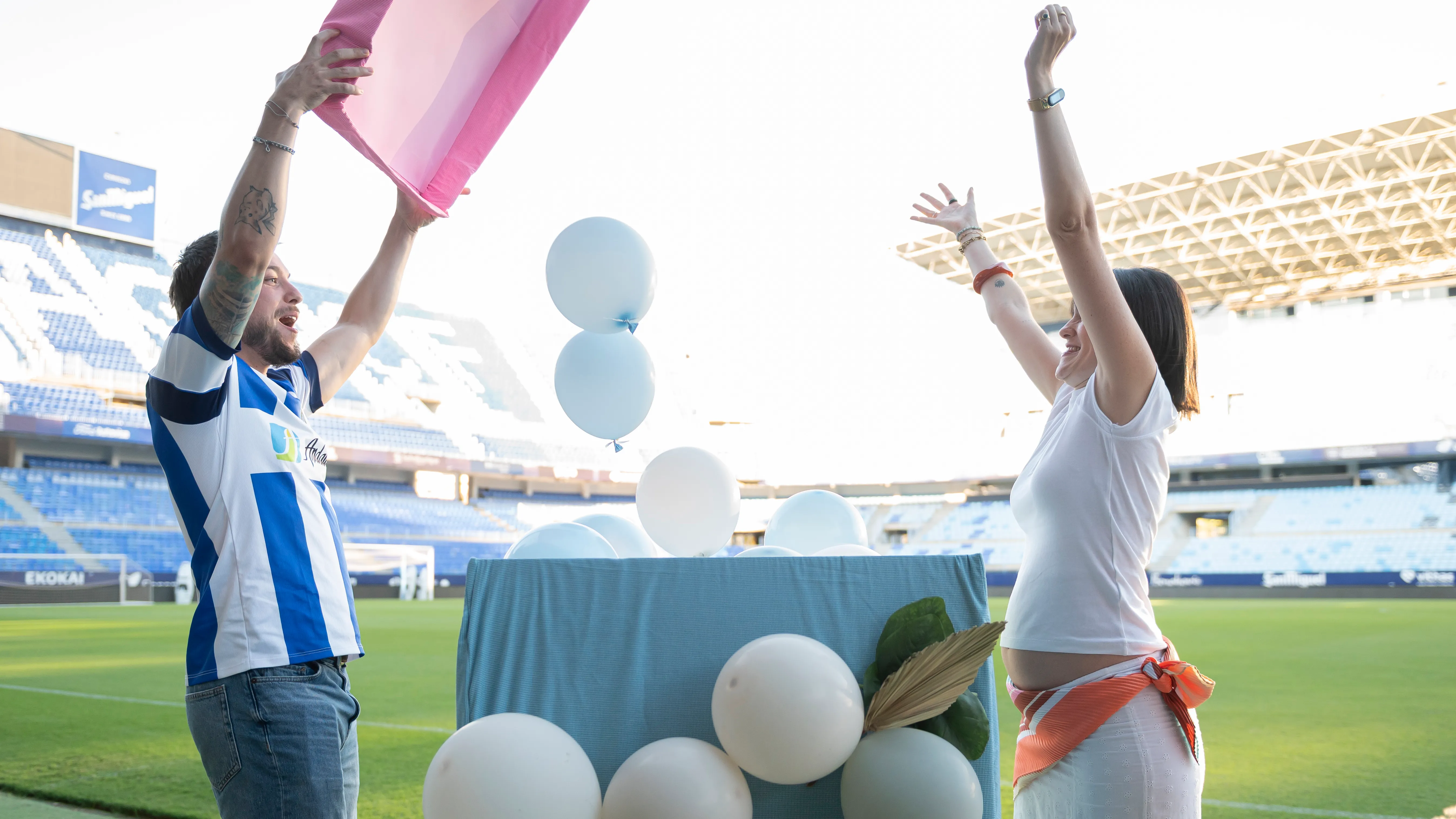 Dos personas celebran con globos blancos en el césped del estadio La Rosaleda; el hombre viste la camiseta blanquiazul del Málaga CF y de fondo se ven las gradas del estadio.
