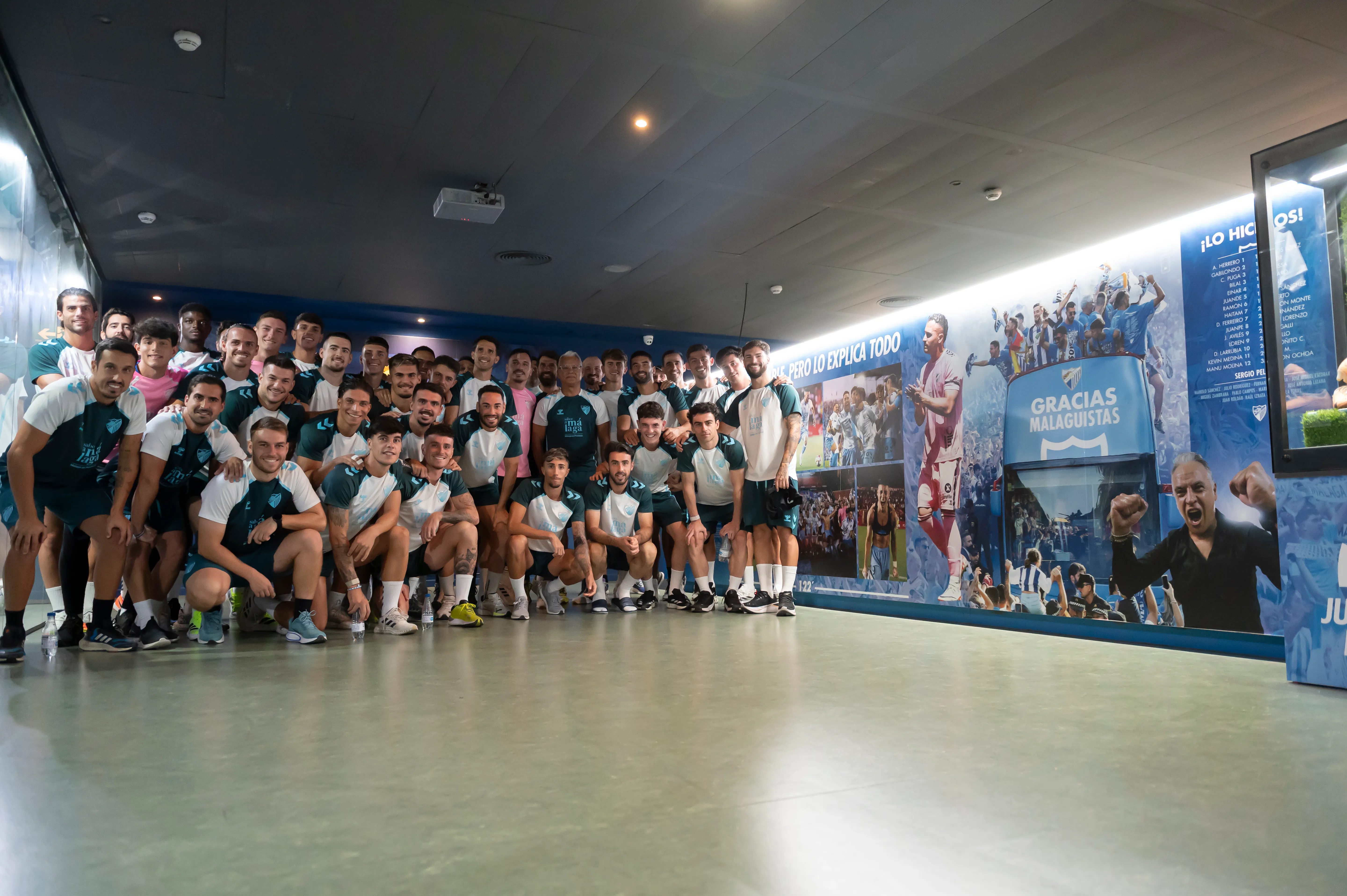 Un gran grupo de jugadores de fútbol y personal posando para una foto dentro de la sala de exposiciones del Museo & Tour Málaga CF, con murales azules que muestran la historia del club y el texto GRACIAS MALAGUISTAS.