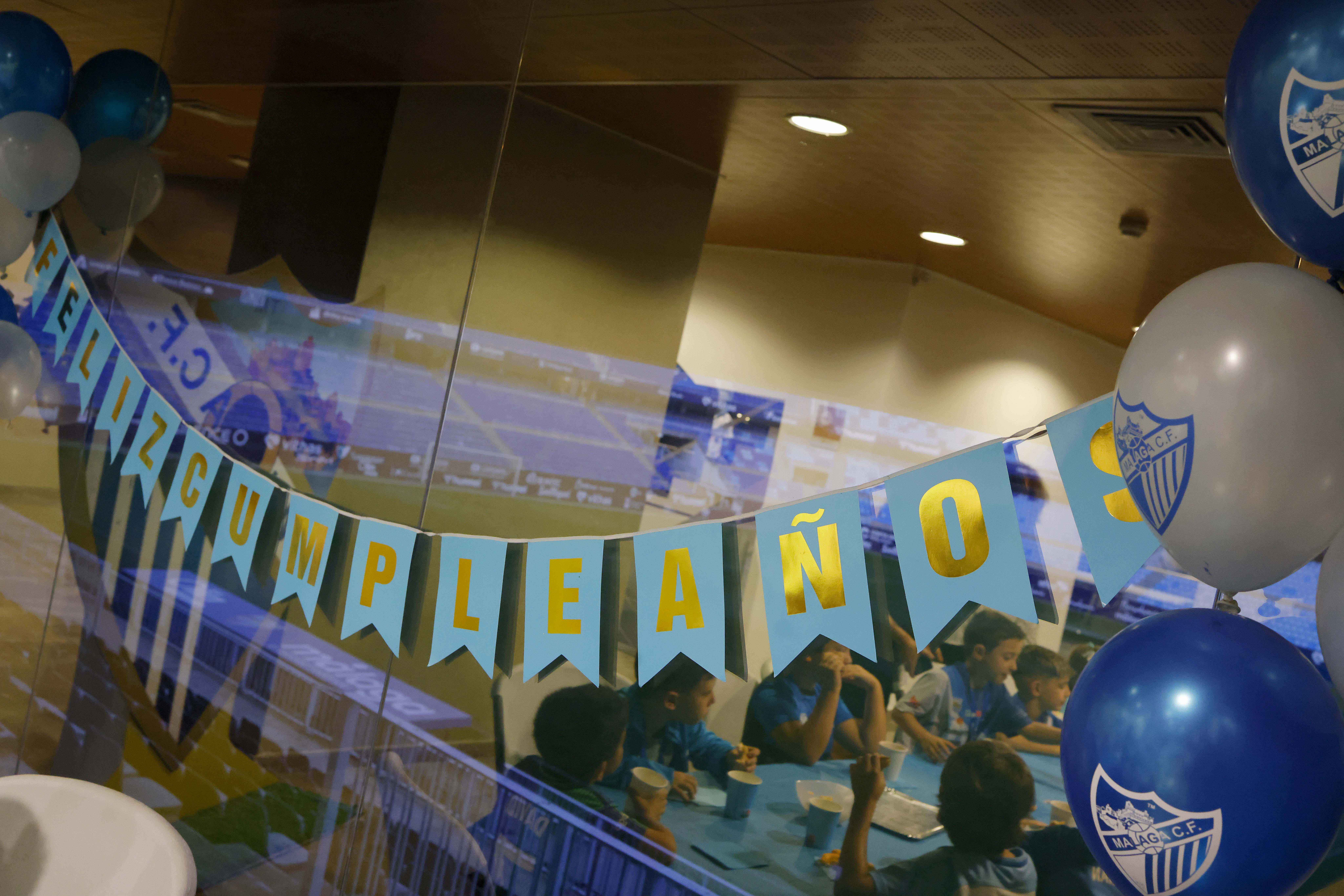 Decoración de fiesta de cumpleaños en interior con globos del Málaga CF en azul y blanco y una pancarta de 'Feliz Cumpleaños', con niños sentados a una mesa al fondo
