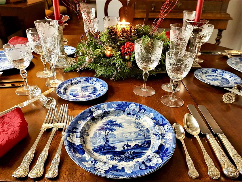 Una mesa de comedor formalmente puesta con platos decorativos azules y blancos, copas de vino de cristal, cubiertos de plata ornamentados y un centro de mesa festivo de hoja perenne con velas rojas sobre una mesa de madera.